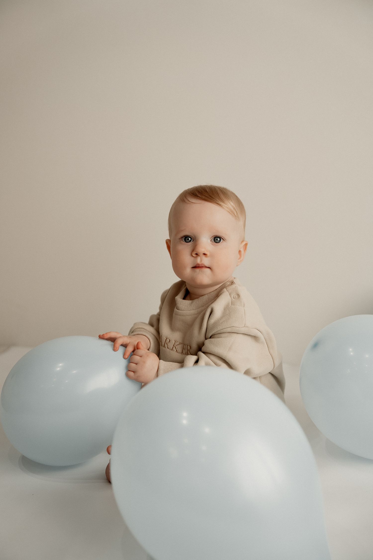 A baby in beige clothing sits on a white surface with light blue balloons arranged around them.