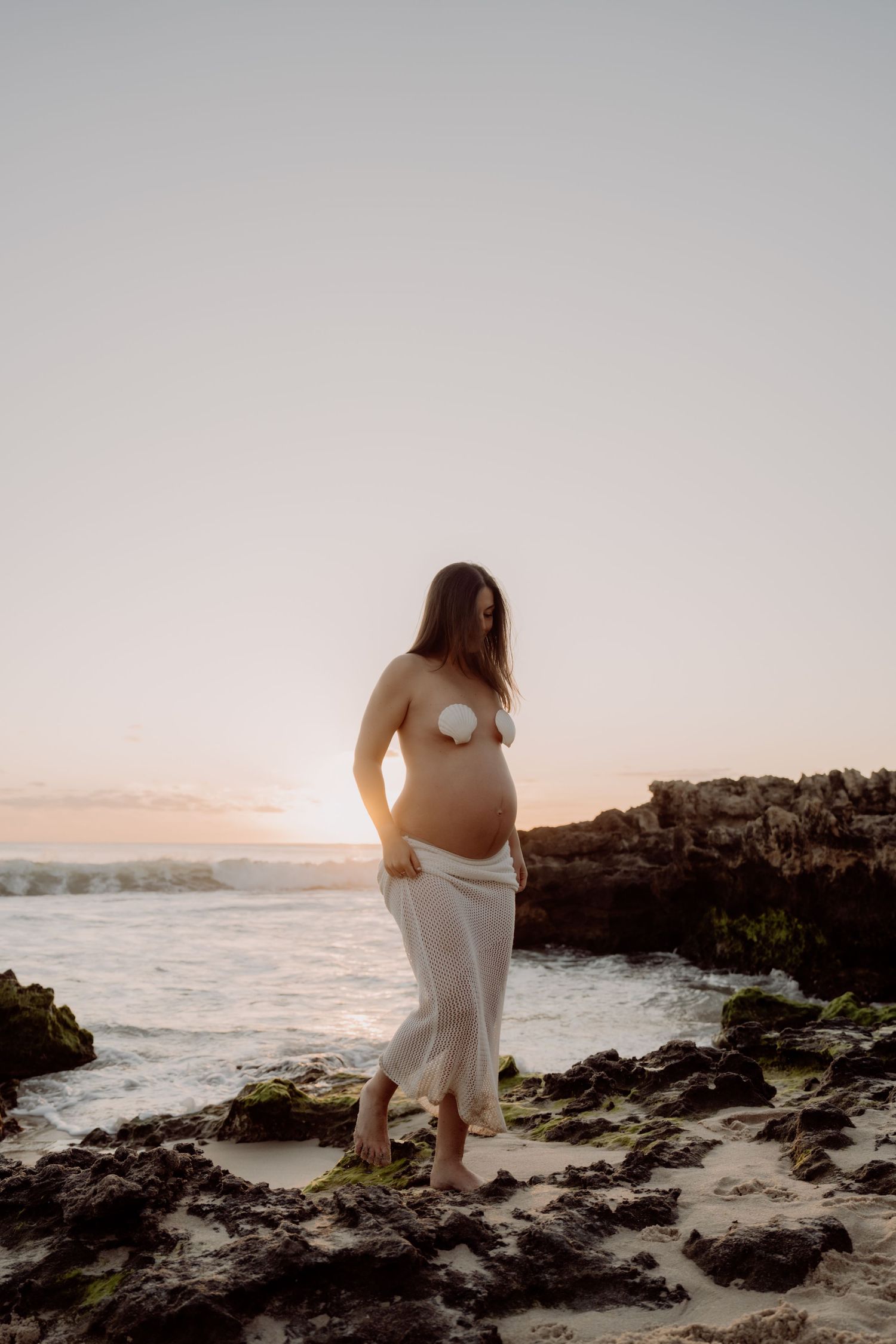 Pregnant woman in white skirt and shell bra standing on rocky beach at sunset with ocean waves behind her.