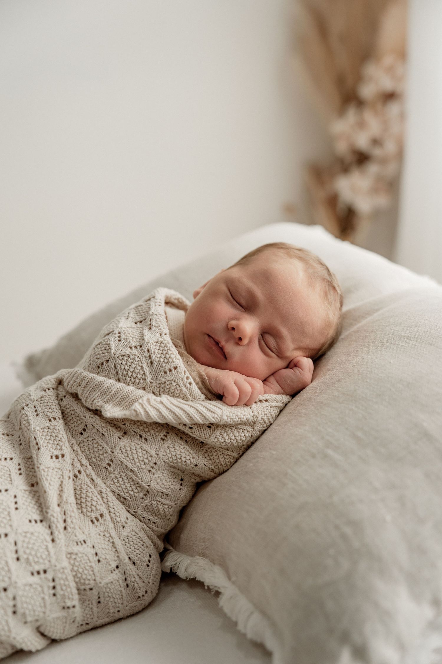 A sleeping newborn baby wrapped in a cream colored knit blanket rests peacefully on white bedding.