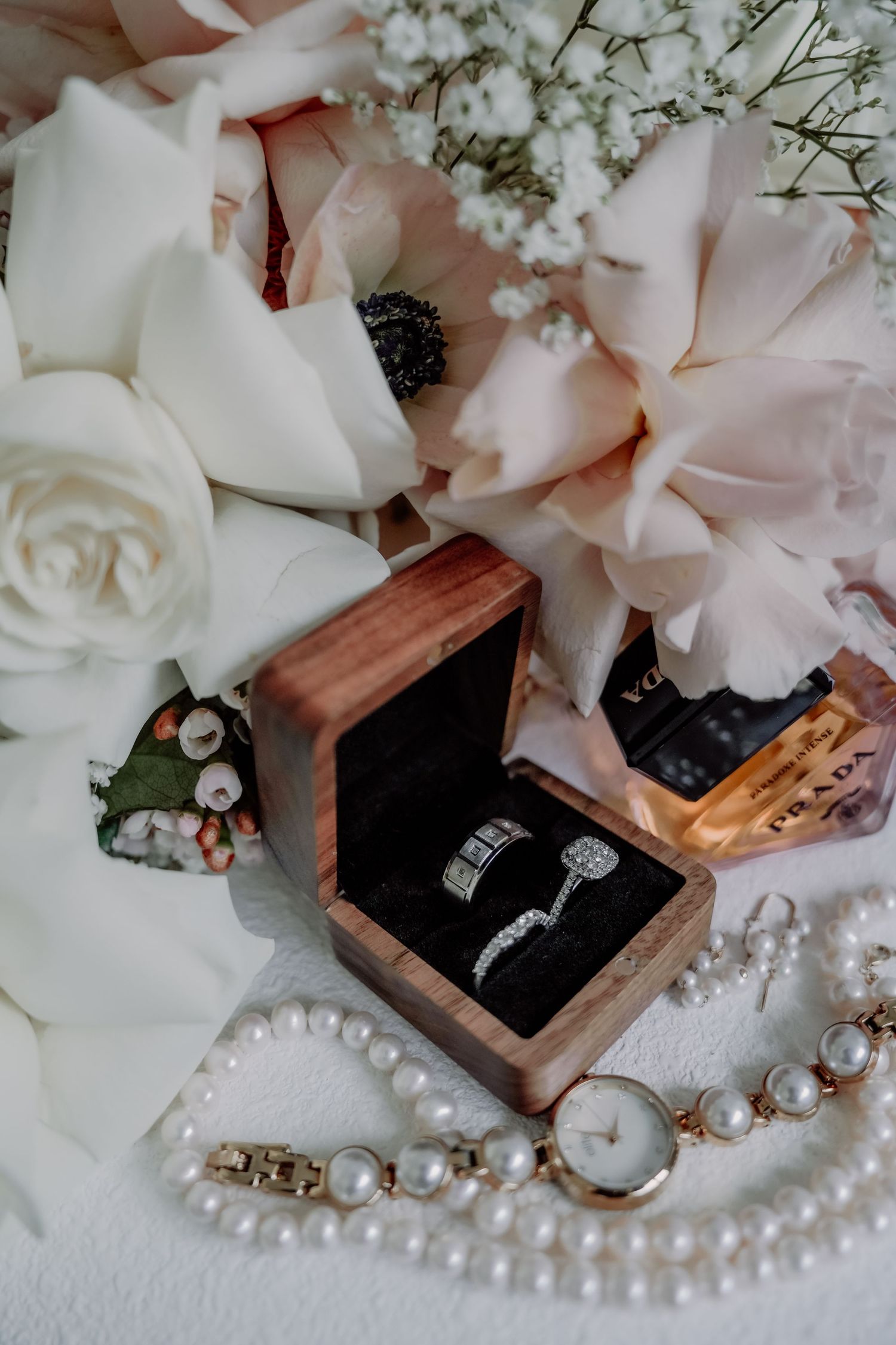 Wedding rings in wooden box surrounded by white roses, baby's breath flowers, and pearl jewelry on white surface.