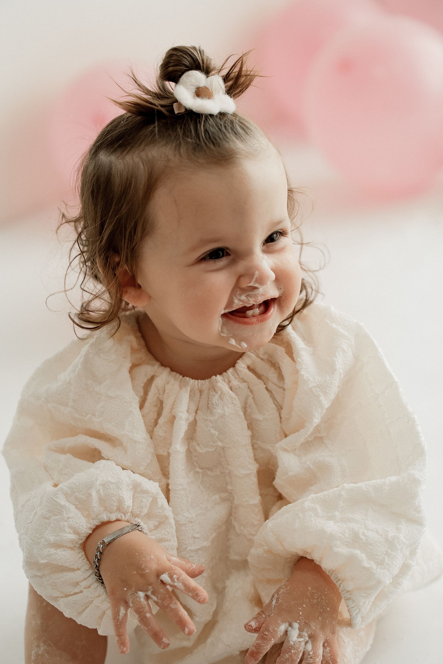 Smiling toddler in white lace dress with hair accessory poses against pink background during portrait session.