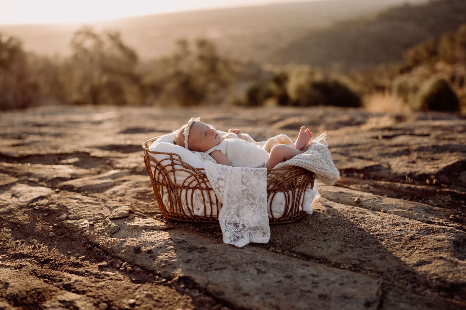 A small infant rests peacefully in a woven basket on a rocky terrain at golden hour.
