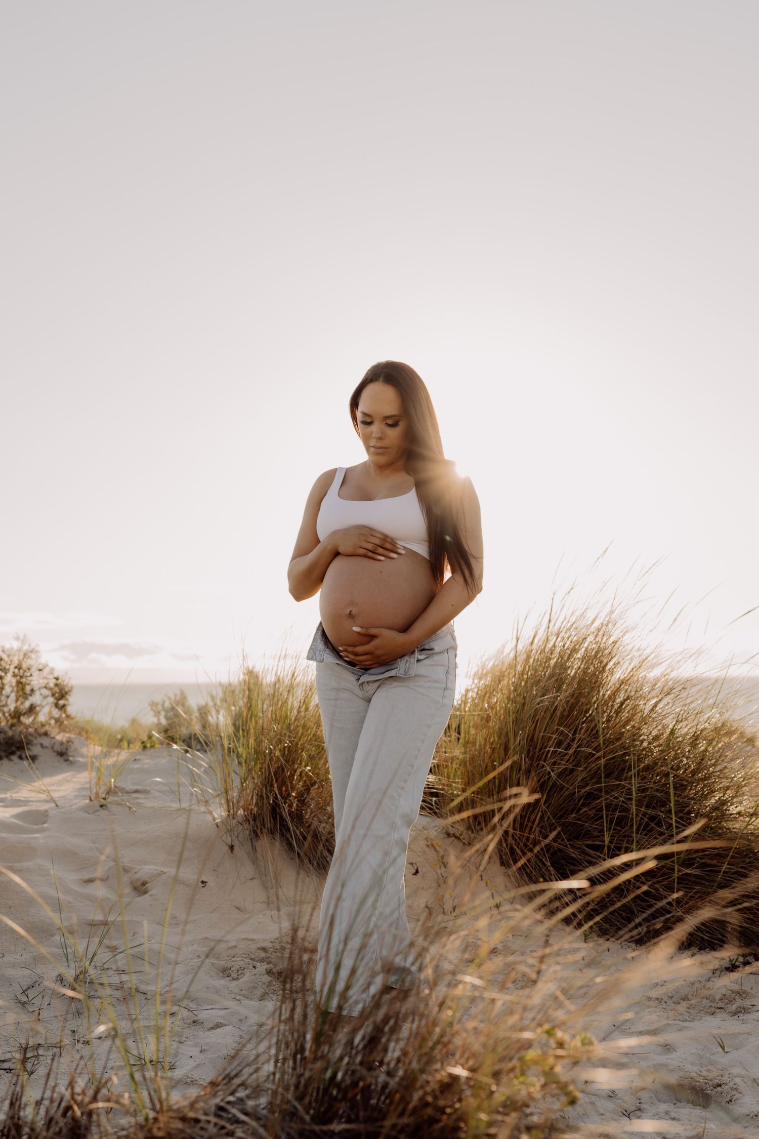A person in a light dress poses on sandy dunes with beach grass at sunset, creating a serene maternity photoshoot scene.