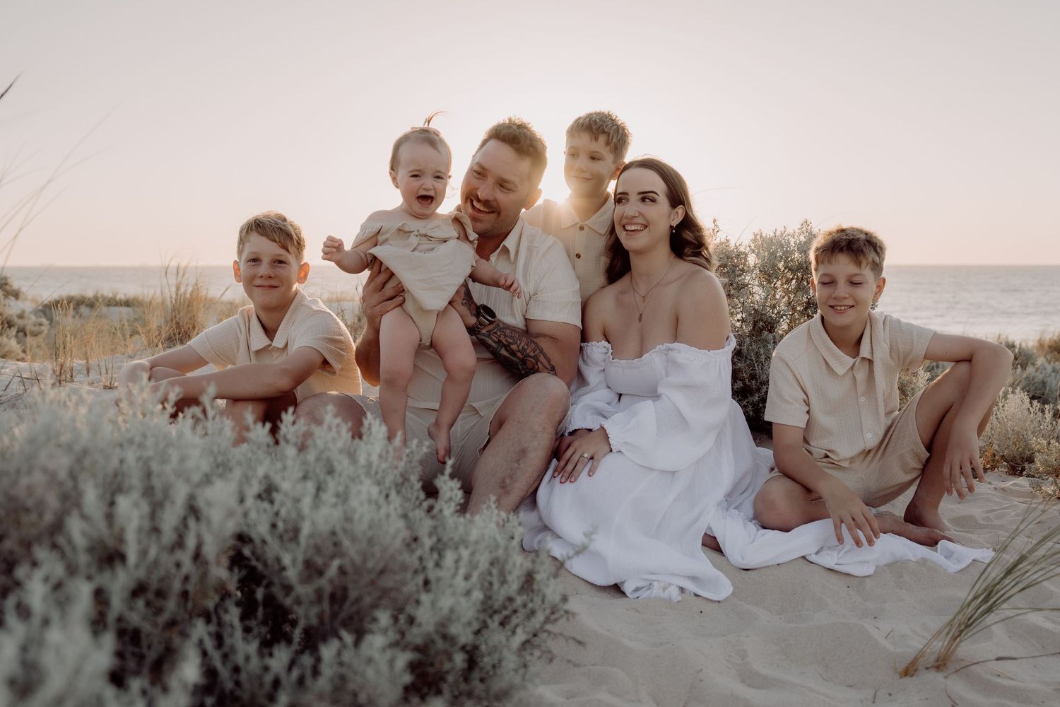 A group sits together in casual attire on sandy ground with desert brush at sunset.