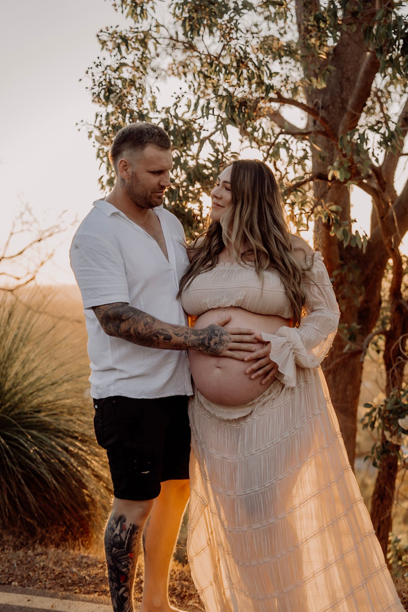 Expecting couple poses together at sunset with the woman wearing a flowing cream lace dress against eucalyptus trees.