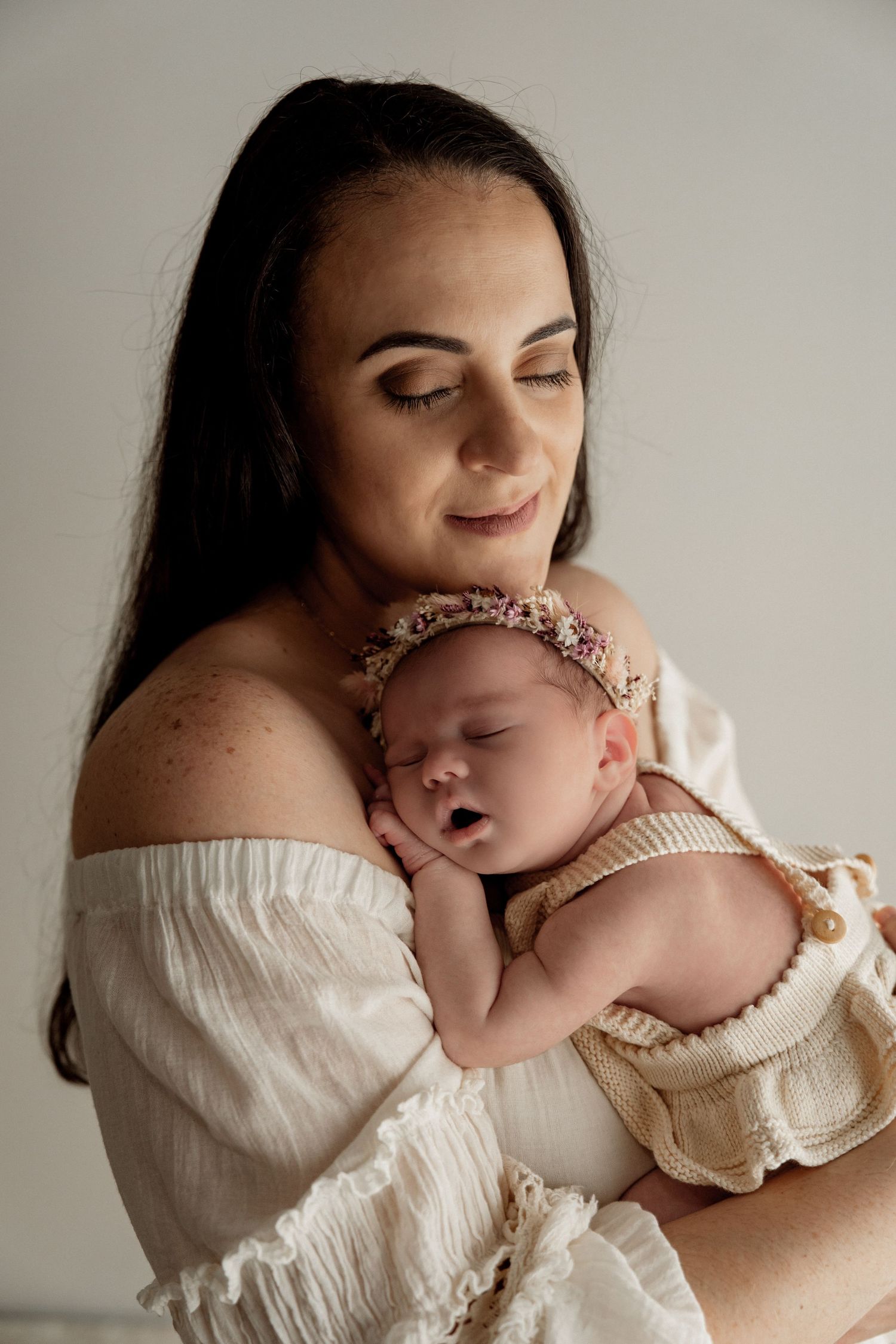 A mother in white peasant blouse holds and cuddles her baby in a tender moment against neutral background.