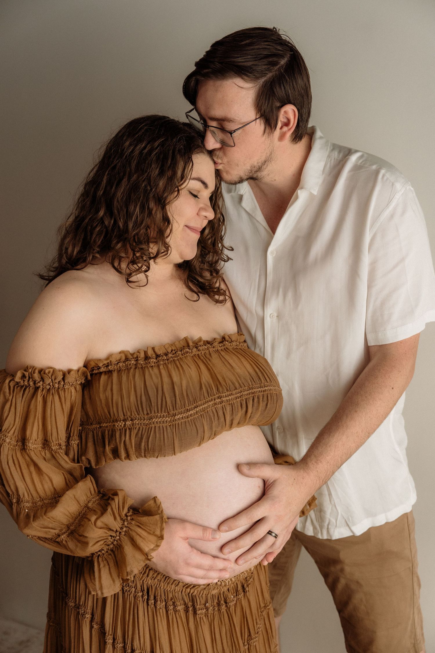Intimate maternity portrait featuring a couple in soft lighting with a brown off-shoulder dress and white shirt.