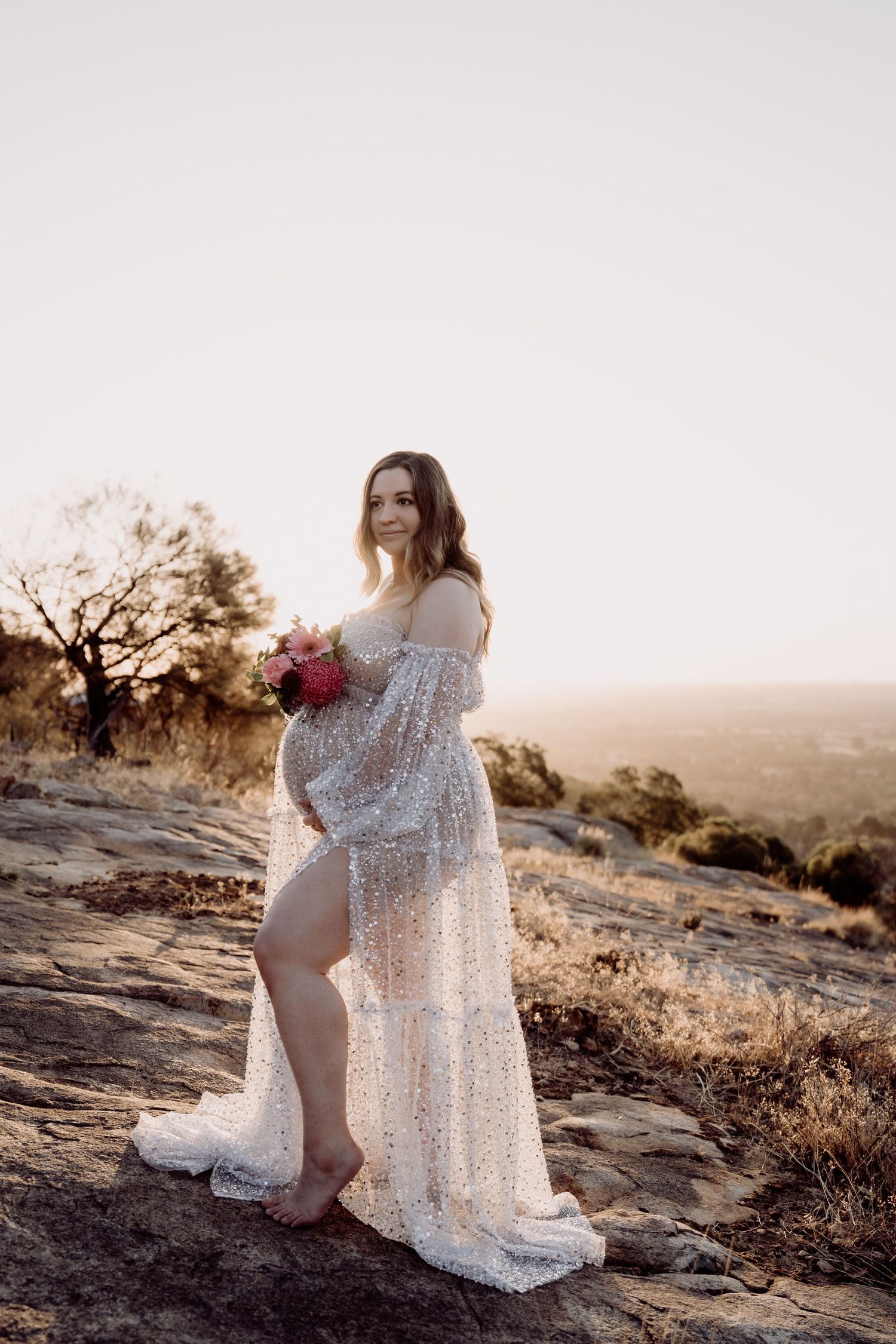 Expectant mother in lace dress poses on rocky outcrop at sunset.