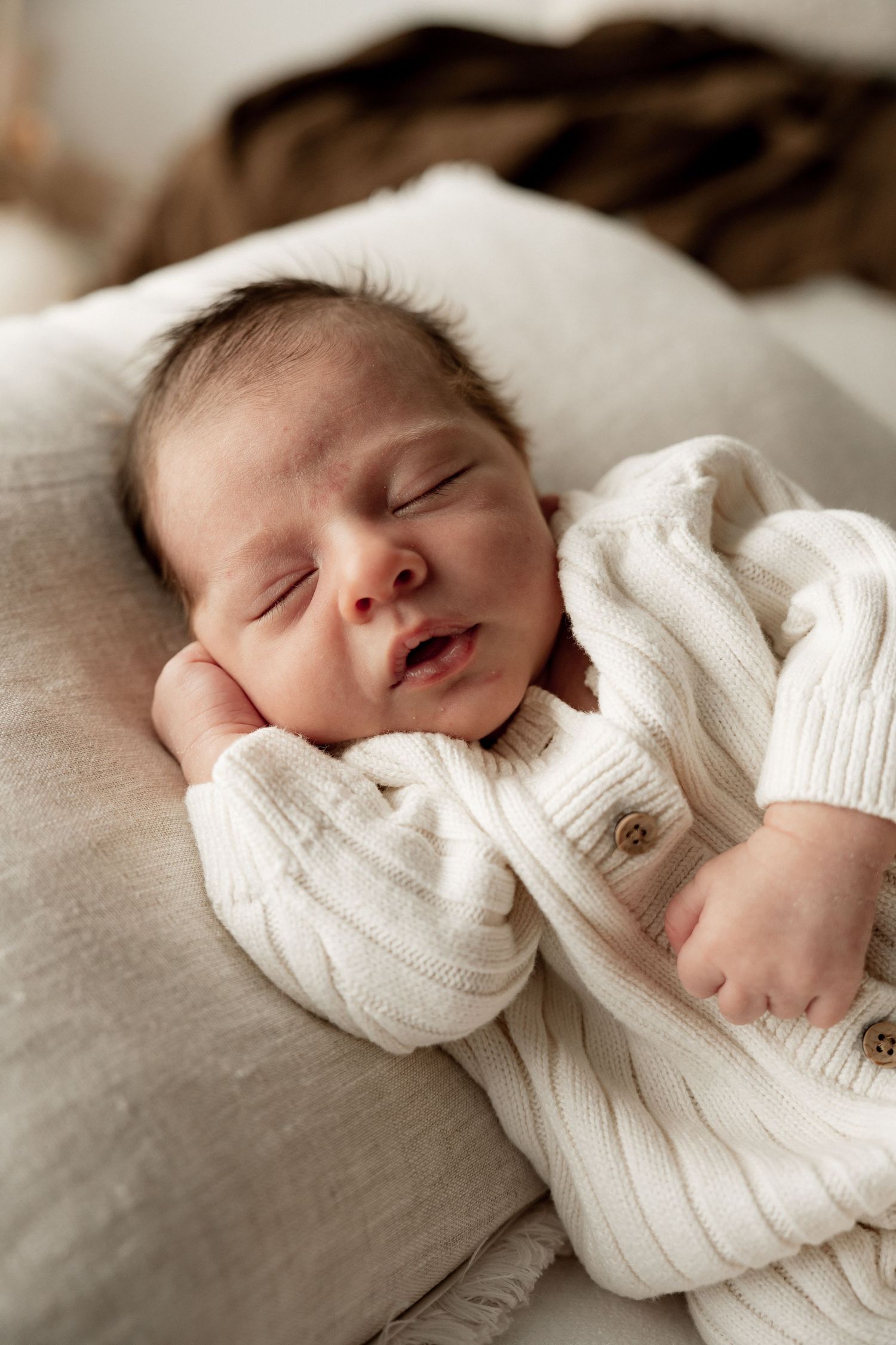 Sleeping newborn baby in white knit outfit resting peacefully on a soft linen surface.