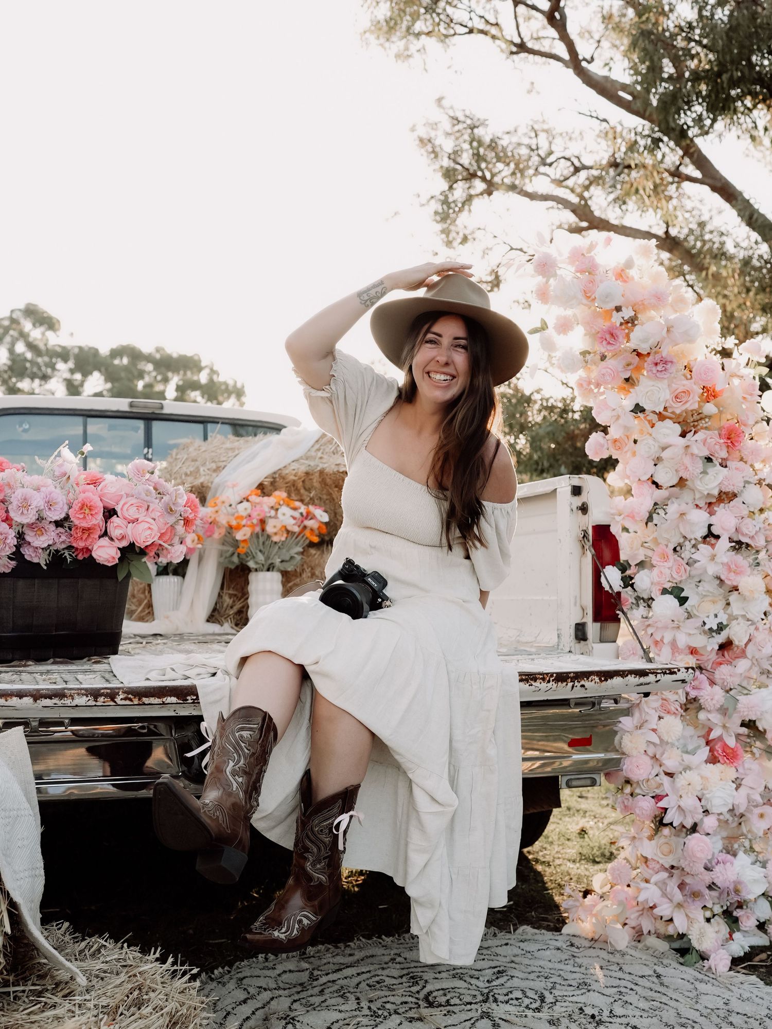 Woman in white dress, cowboy boots, and hat sits on flower-adorned truck bed surrounded by pink blooms outdoors.