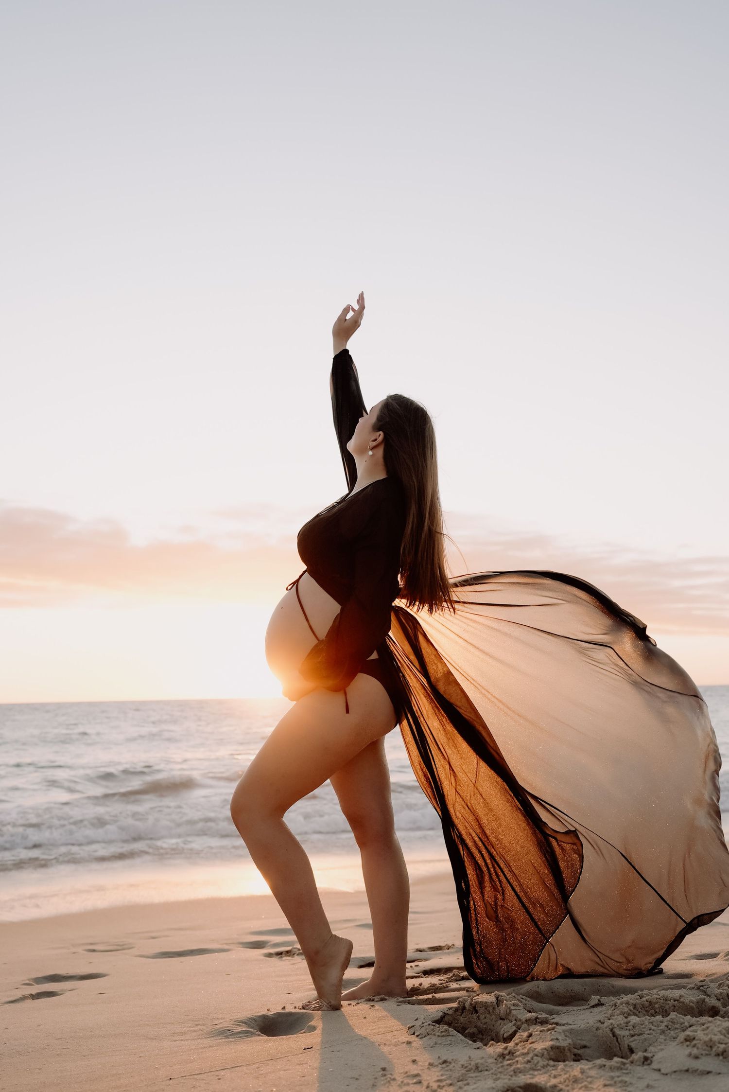 Pregnant woman in black bodysuit with flowing sheer fabric poses gracefully on beach at golden sunset.