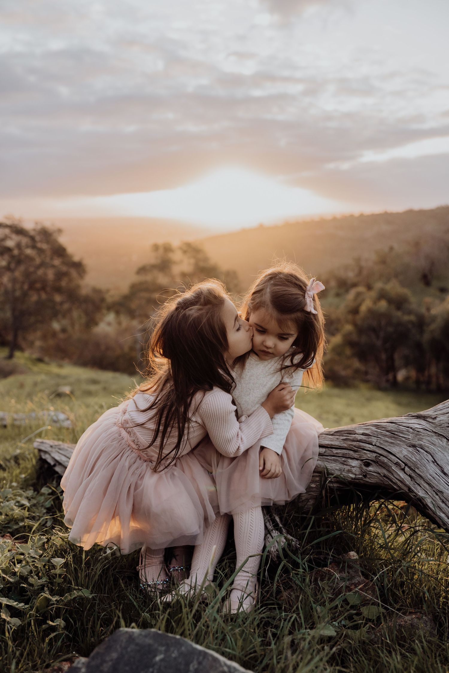 Two young people in white dresses sitting on a fallen log at sunset, sharing a tender moment in nature.