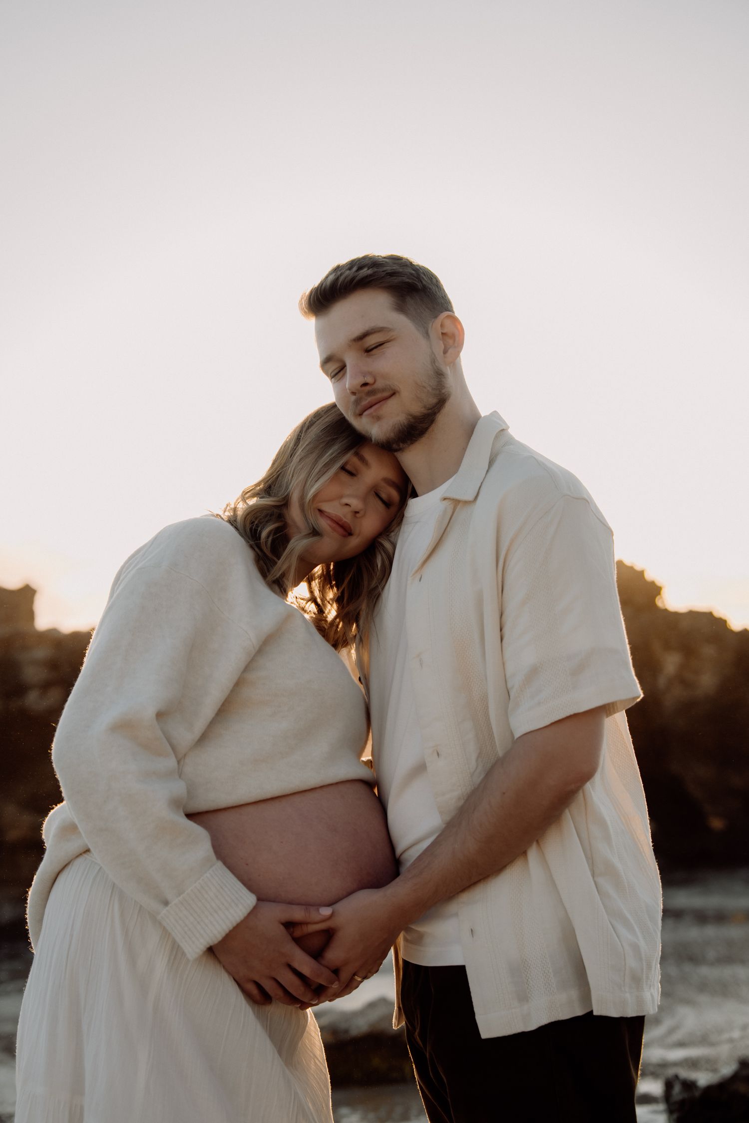 A romantic couple in white clothing embraces tenderly during sunset in an outdoor setting.