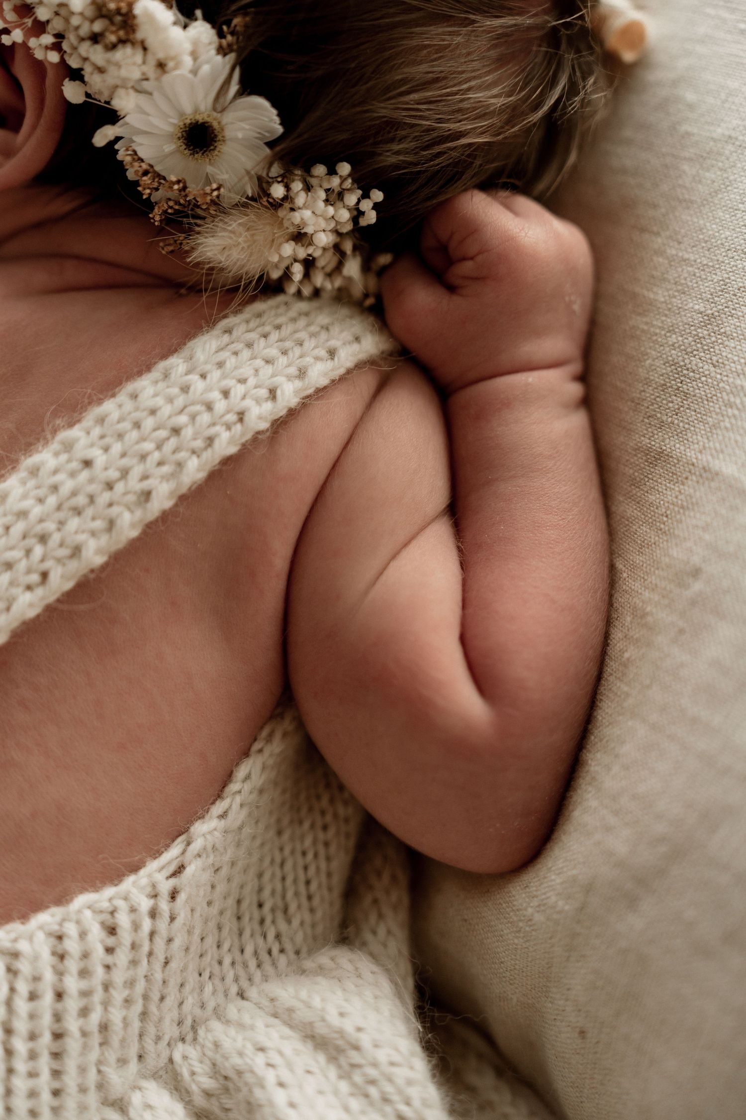 Close-up of a sleeping newborn baby's chubby arm and hand with a floral crown, wrapped in a cream knit blanket.