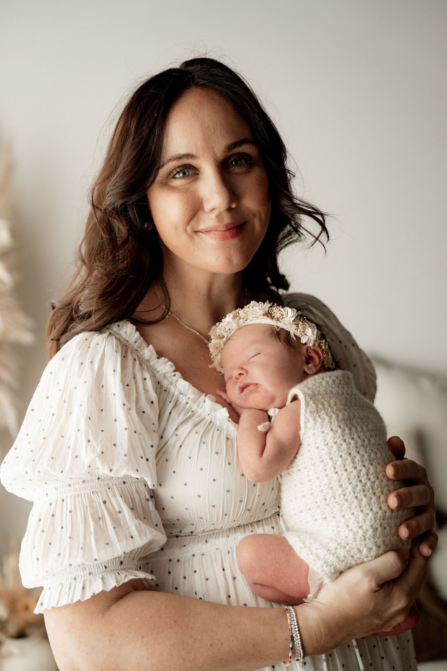 A smiling mother in a white dress tenderly holds her swaddled newborn baby wearing a floral headband.