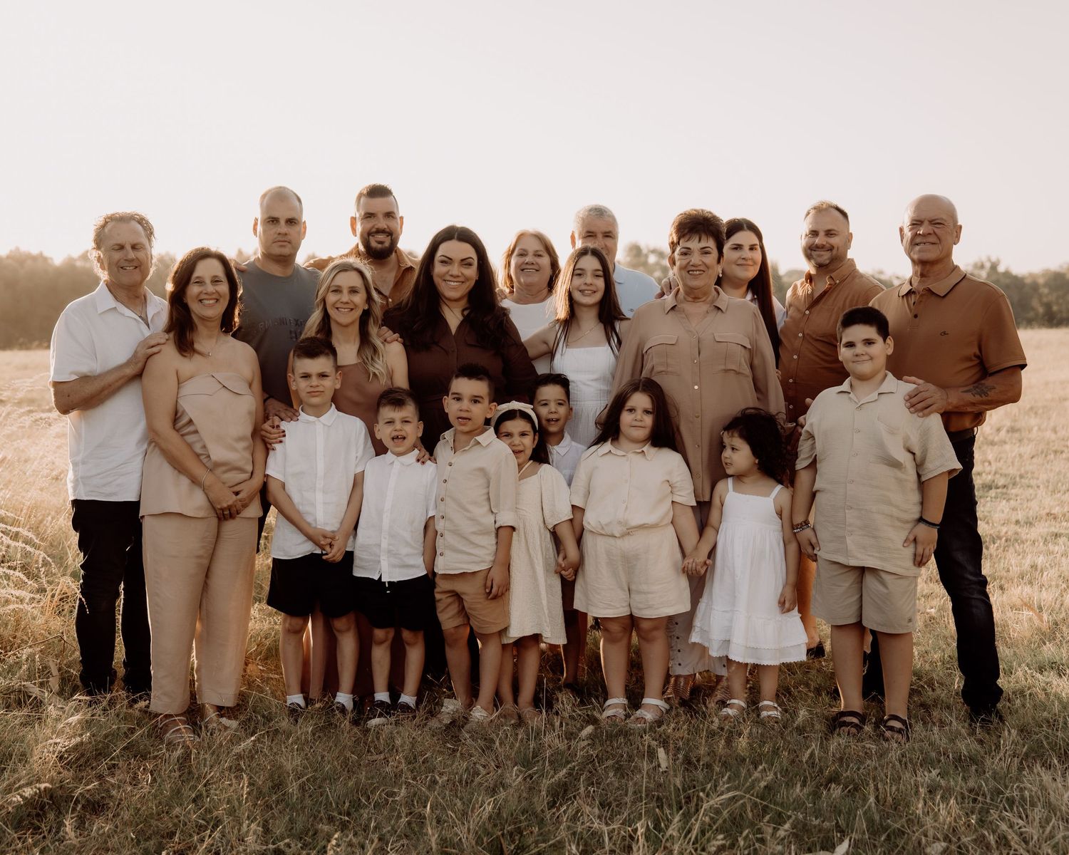 A large family group in coordinated beige and white outfits poses together in a sunlit outdoor field setting.