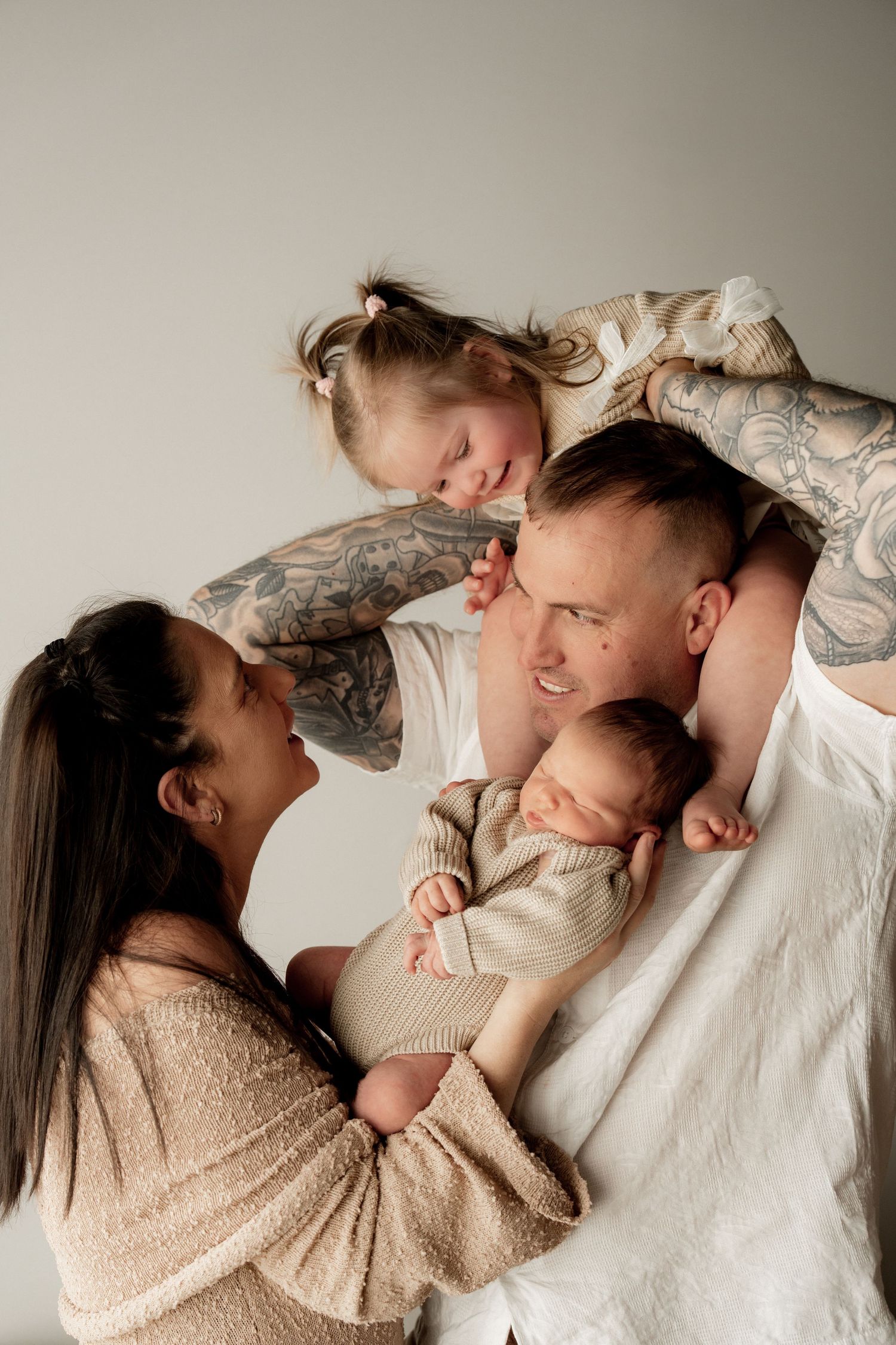 A tattooed couple poses with their newborn baby and young daughter in a warm, intimate family portrait against a gray background.