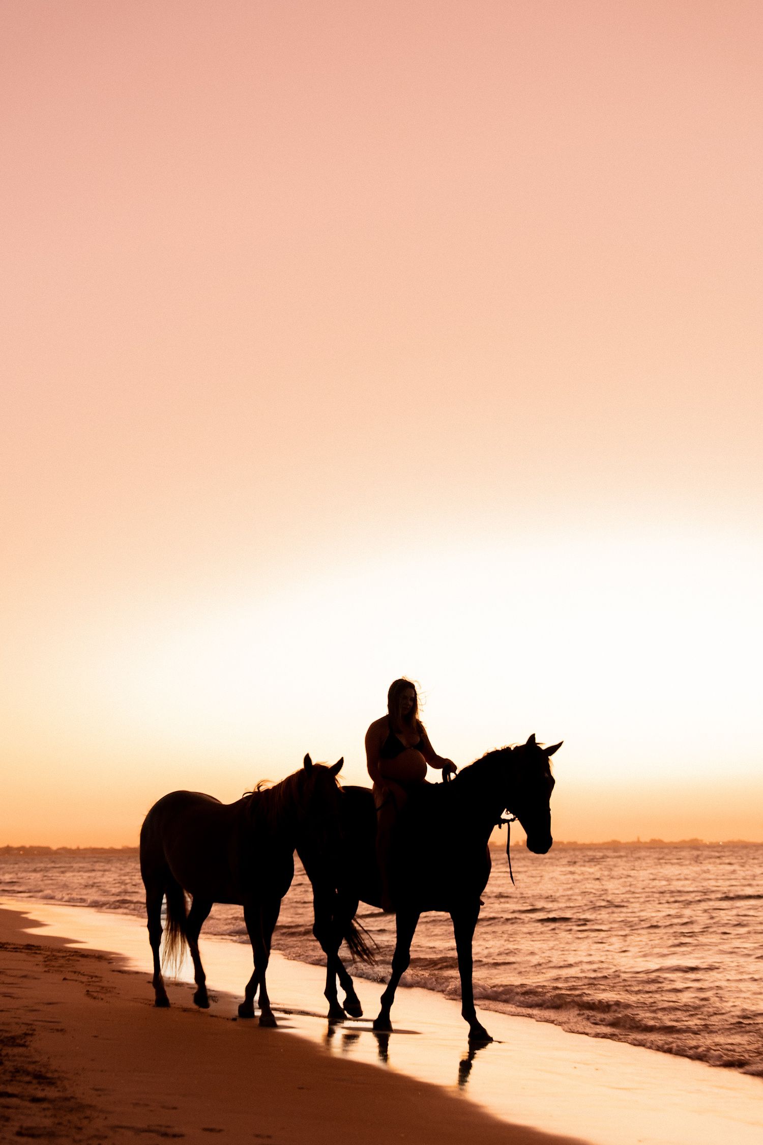 Silhouette of person horseback riding with two horses along a beach at sunset with orange sky and ocean.