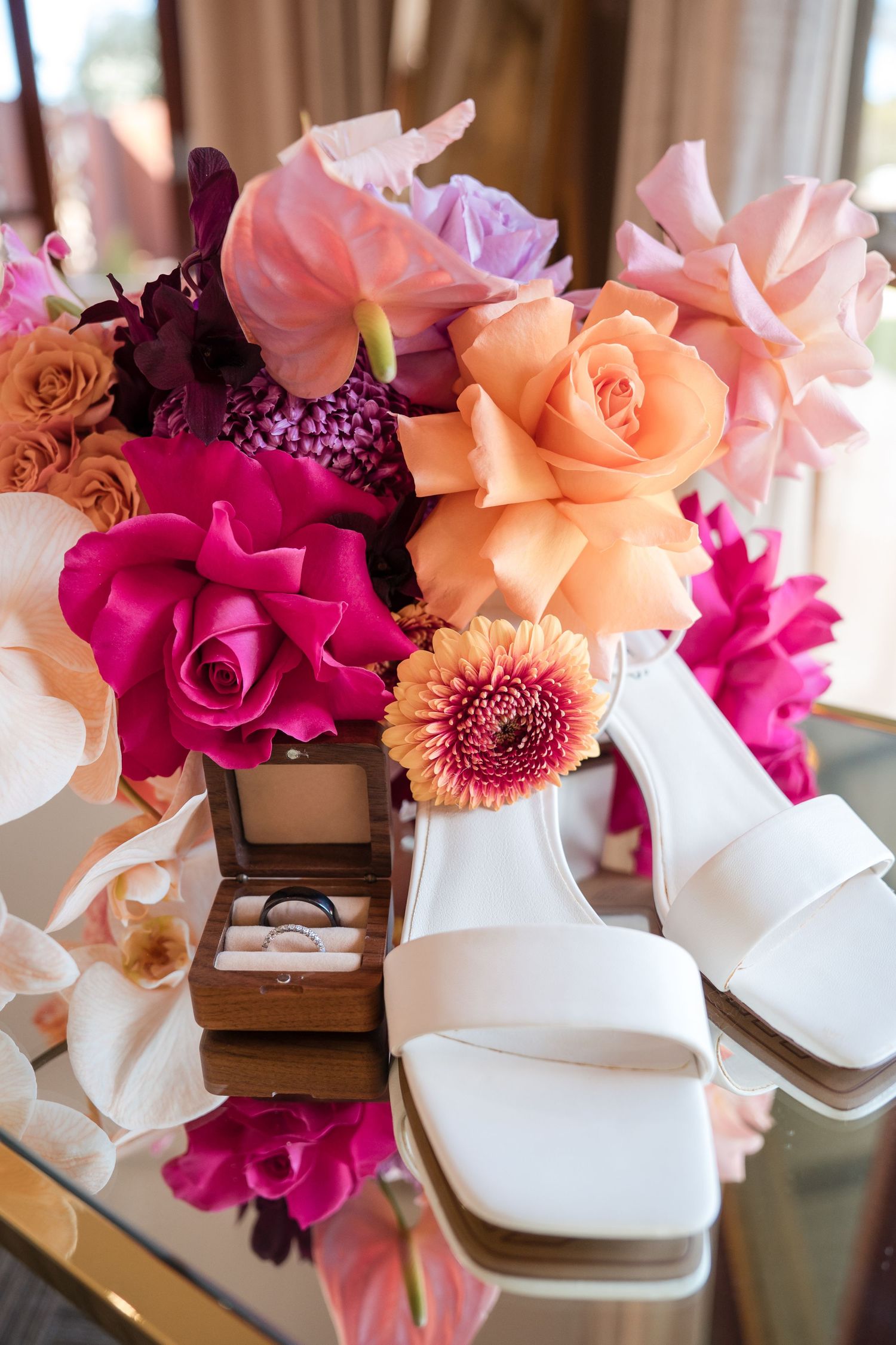 Beautiful pink and peach roses arranged with white orchids next to elegant white bridal shoes on a wooden surface.