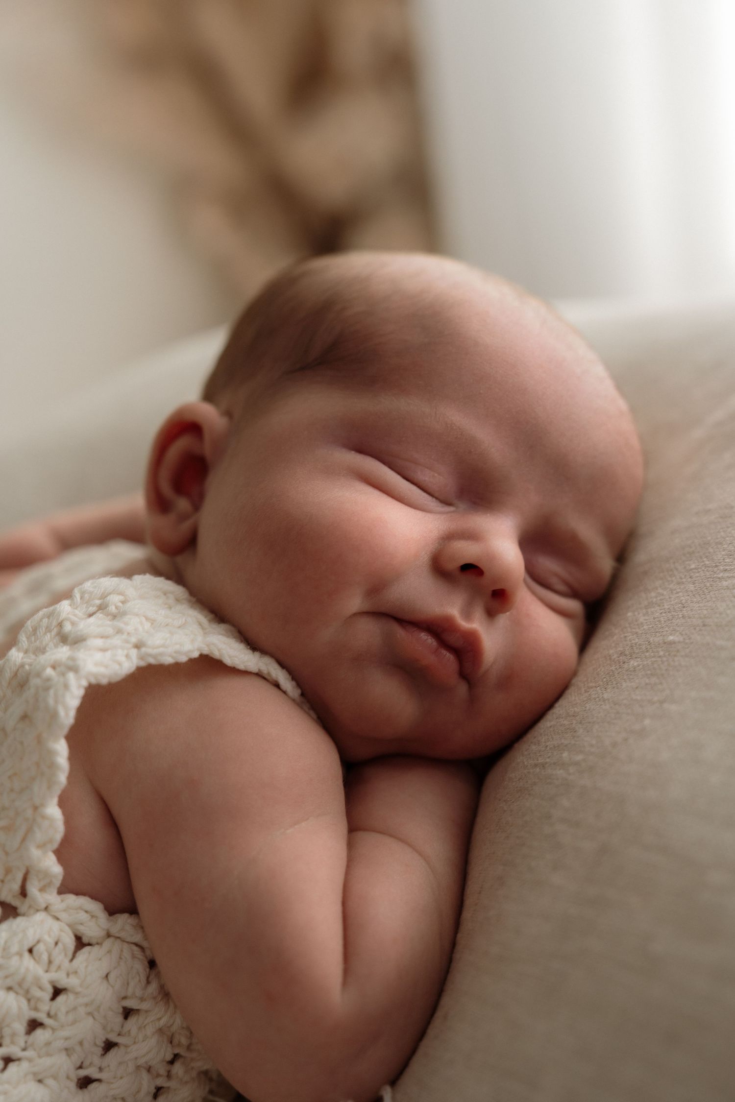 Sleeping newborn wearing white lace outfit rests peacefully on soft beige cushion.