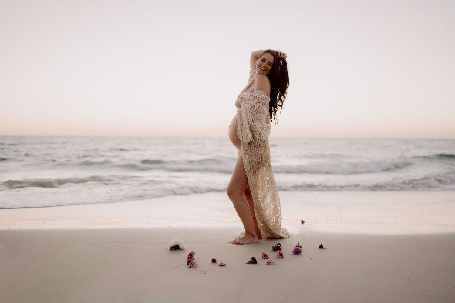 A figure in a flowing white dress walks gracefully along a misty beach shoreline at sunset.