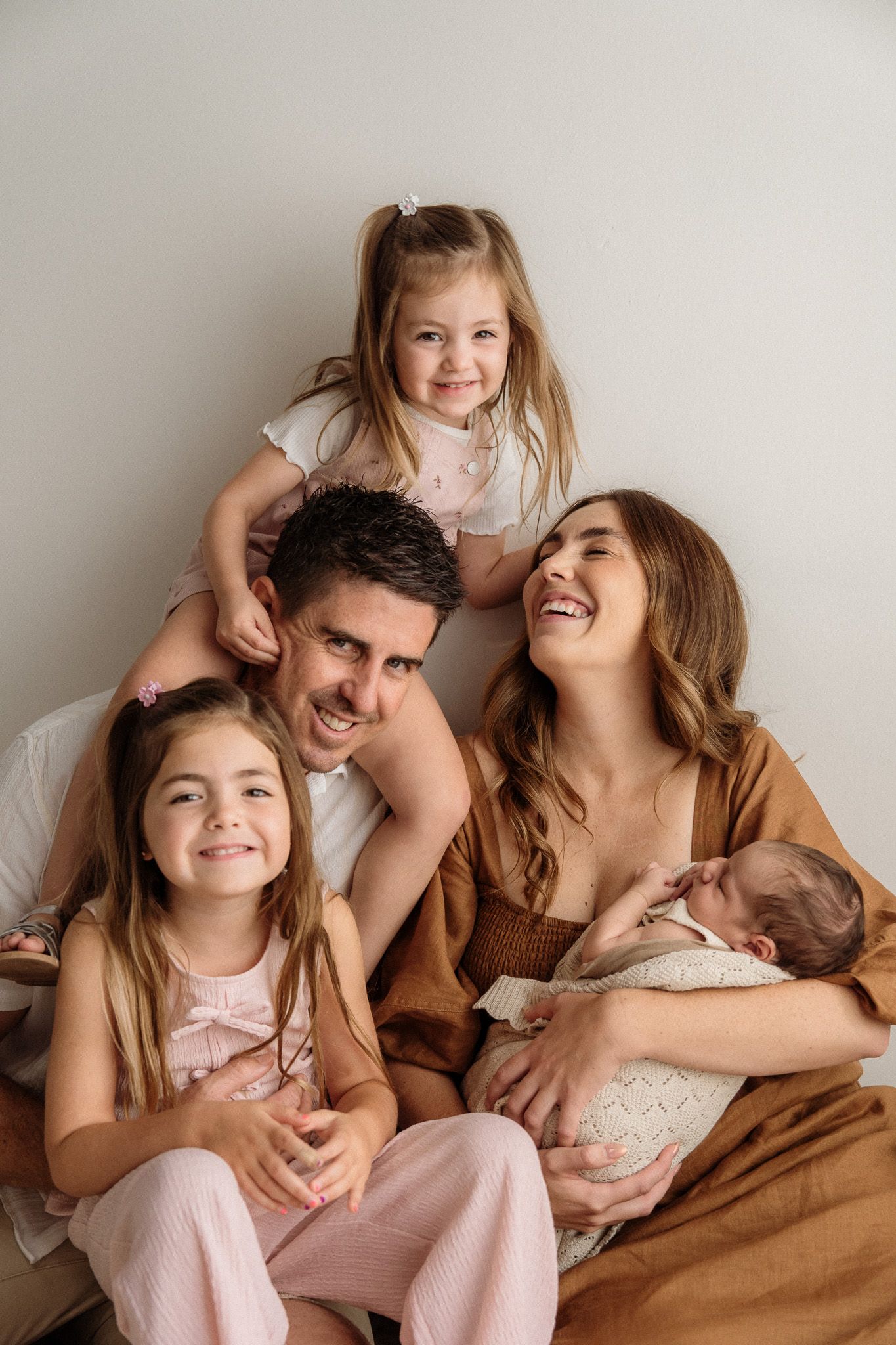 A joyful family cuddles and laughs together while wearing cozy brown and pink outfits against a neutral backdrop.