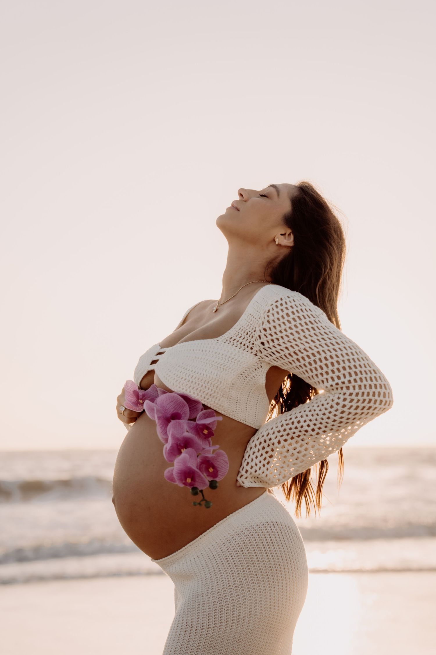 A pregnant figure in a white crochet dress poses at sunset on the beach with purple orchids decorating their belly.
