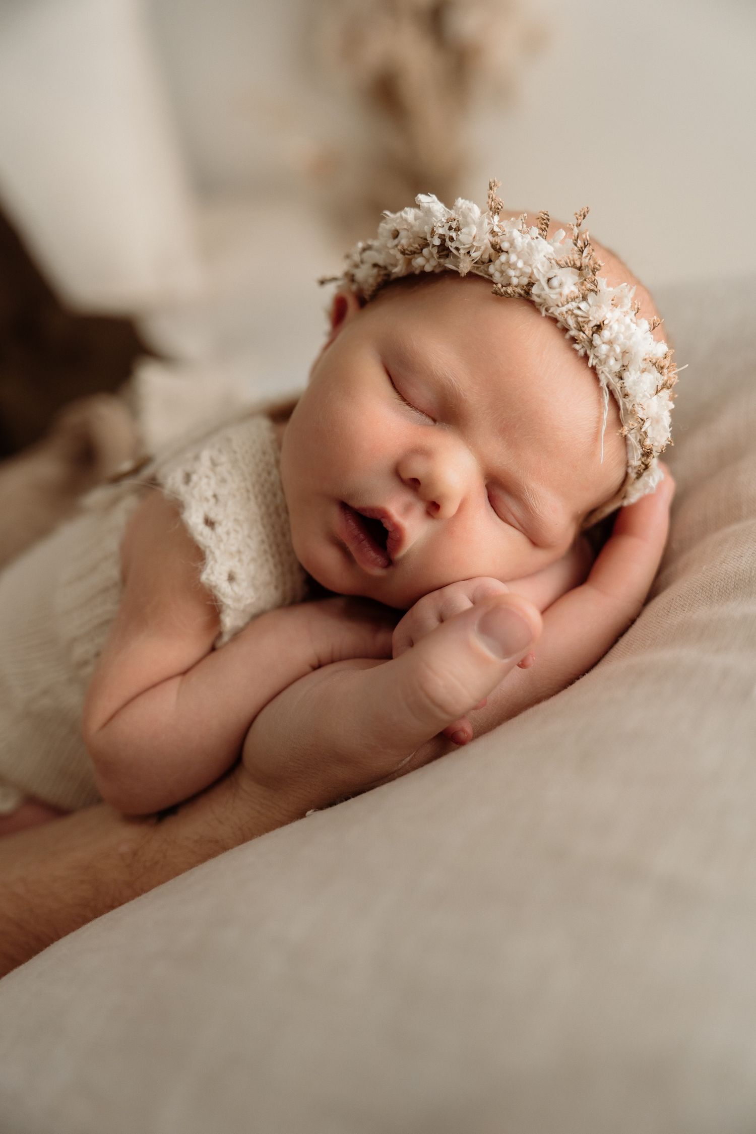 Peaceful sleeping newborn wearing delicate white lace dress and floral crown rests on soft cream bedding.