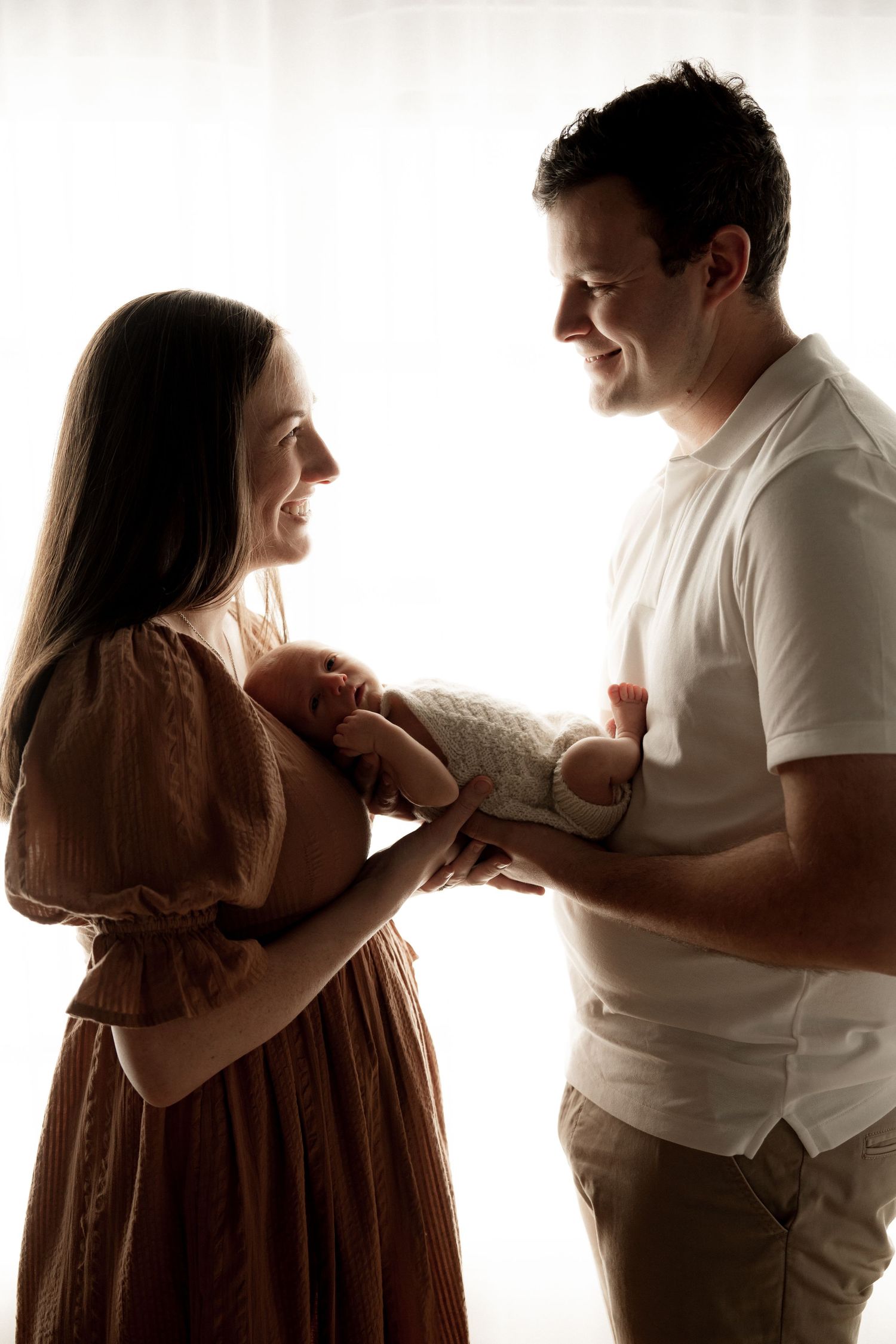 A couple smiles lovingly at their newborn baby, bathed in soft backlit glow, creating a warm family portrait.