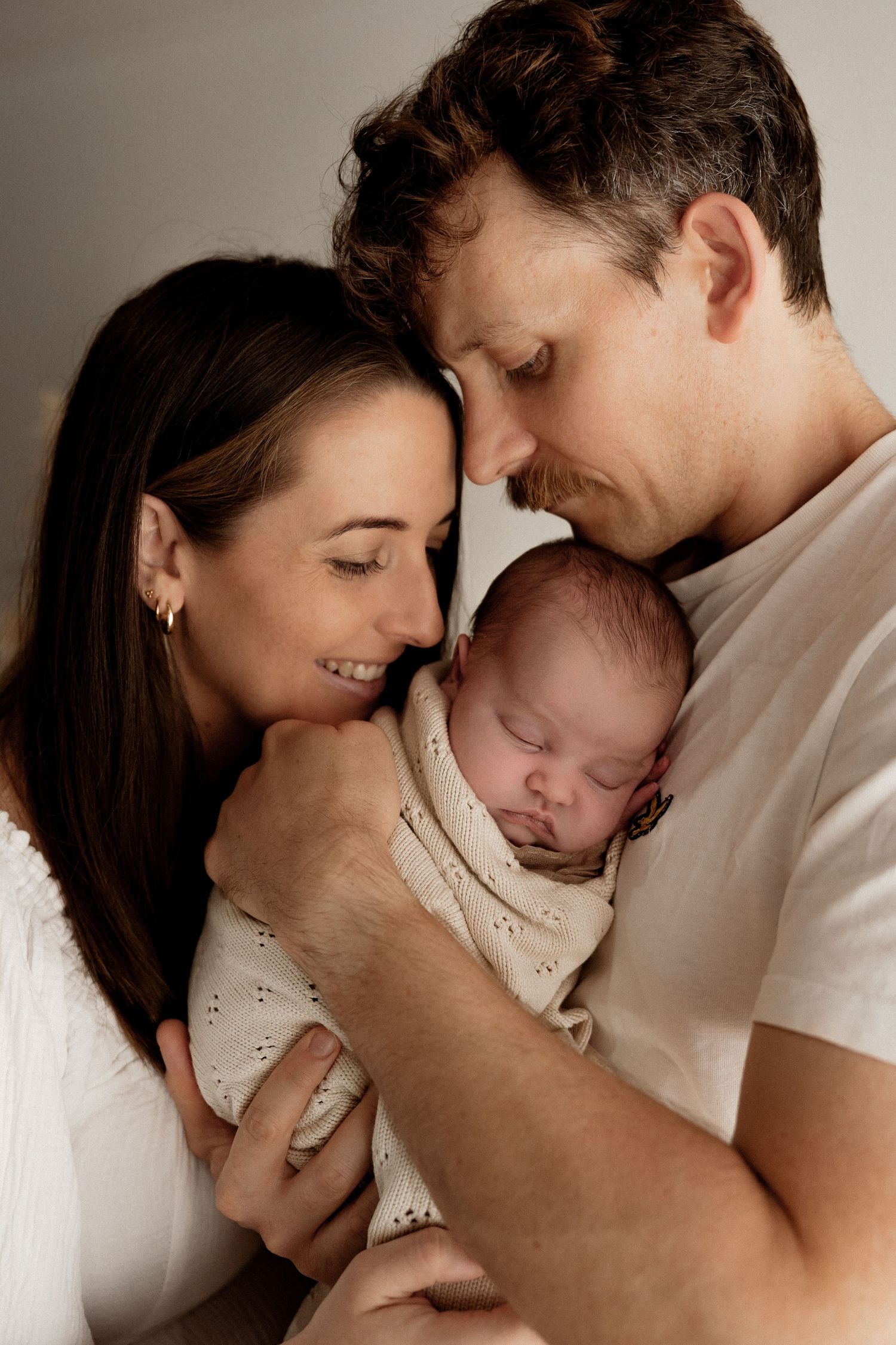 New parents holding and gazing lovingly at their sleeping newborn baby wrapped in a cream-colored dotted outfit.