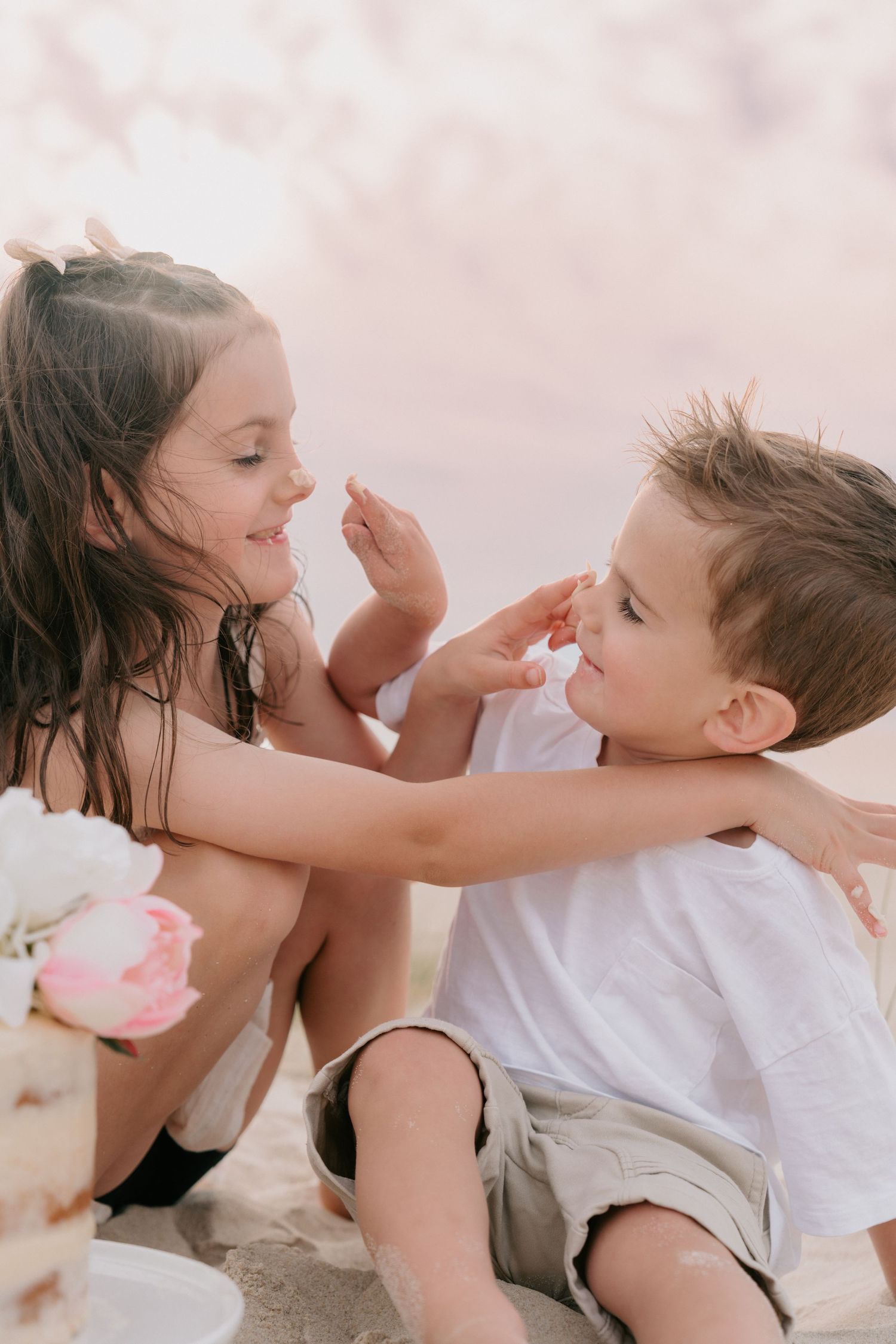 Two young children playfully touching each other's faces while sitting on a sandy beach, sharing a joyful moment.