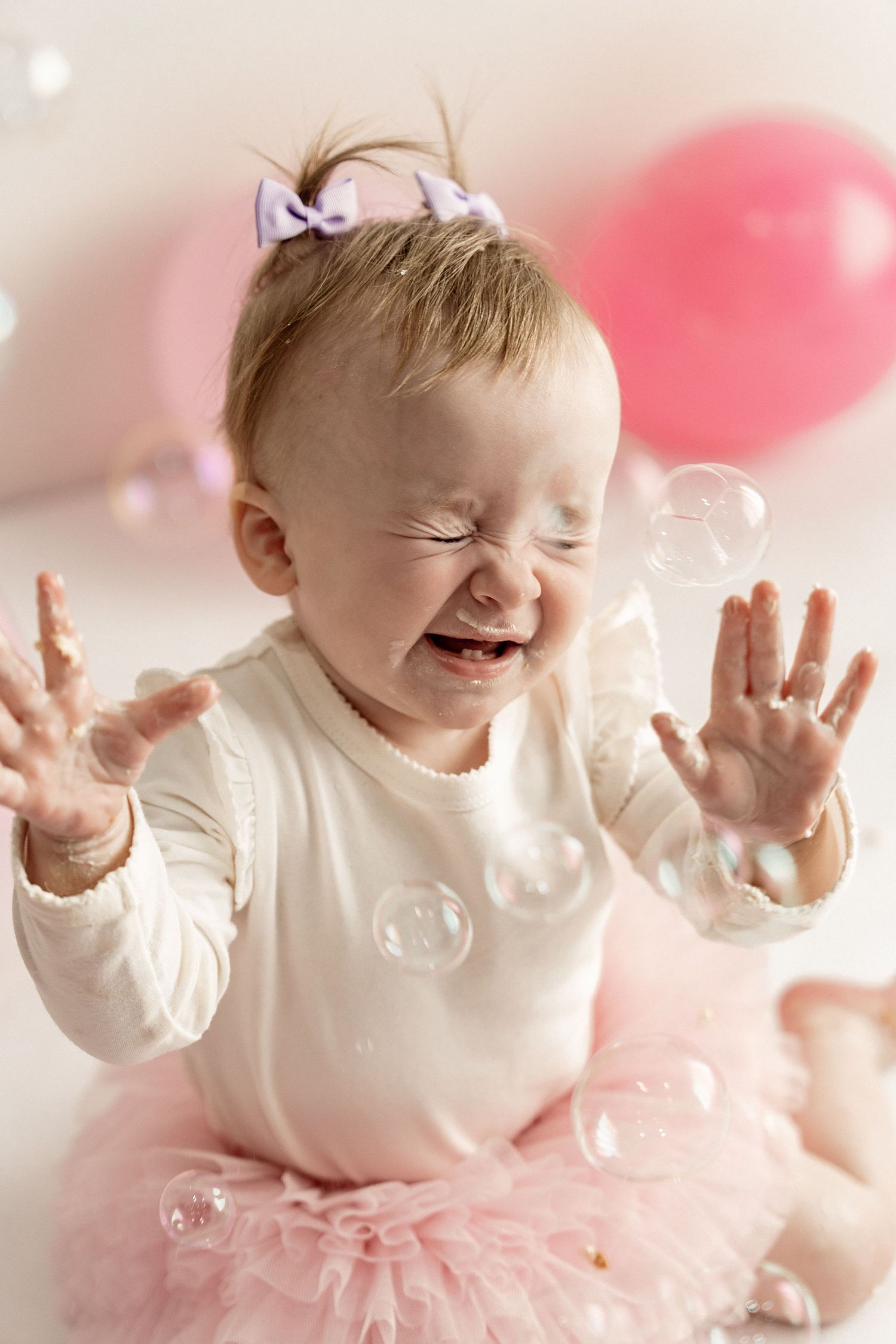 A baby in a white top and pink tutu smiles and plays with soap bubbles against a soft pink background.