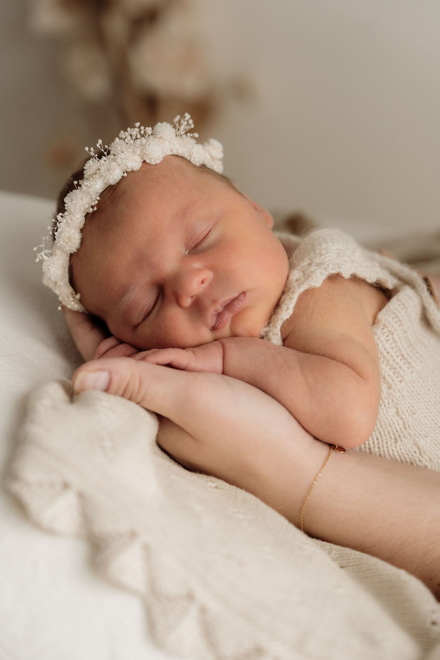 A sleeping newborn baby wearing a white wrap and delicate lace headband rests peacefully on soft white bedding.
