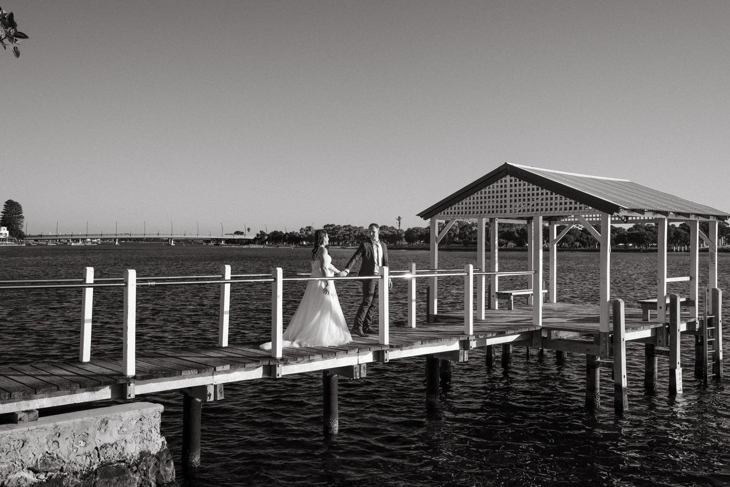 A panoramic black and white photograph shows a long wooden pier stretching into calm waters under an overcast sky.