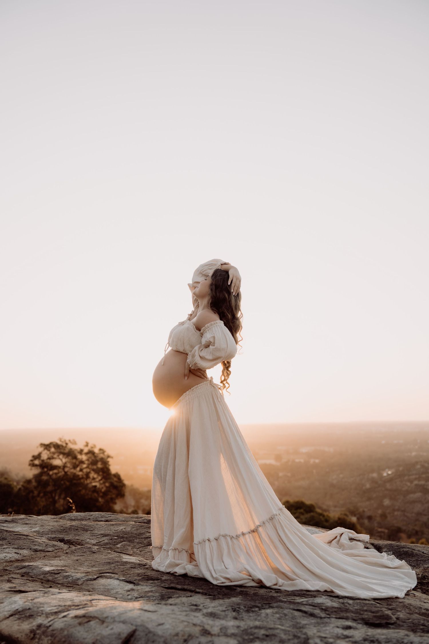 A bride in a flowing white wedding dress with a long train poses against a sunset mountain landscape.