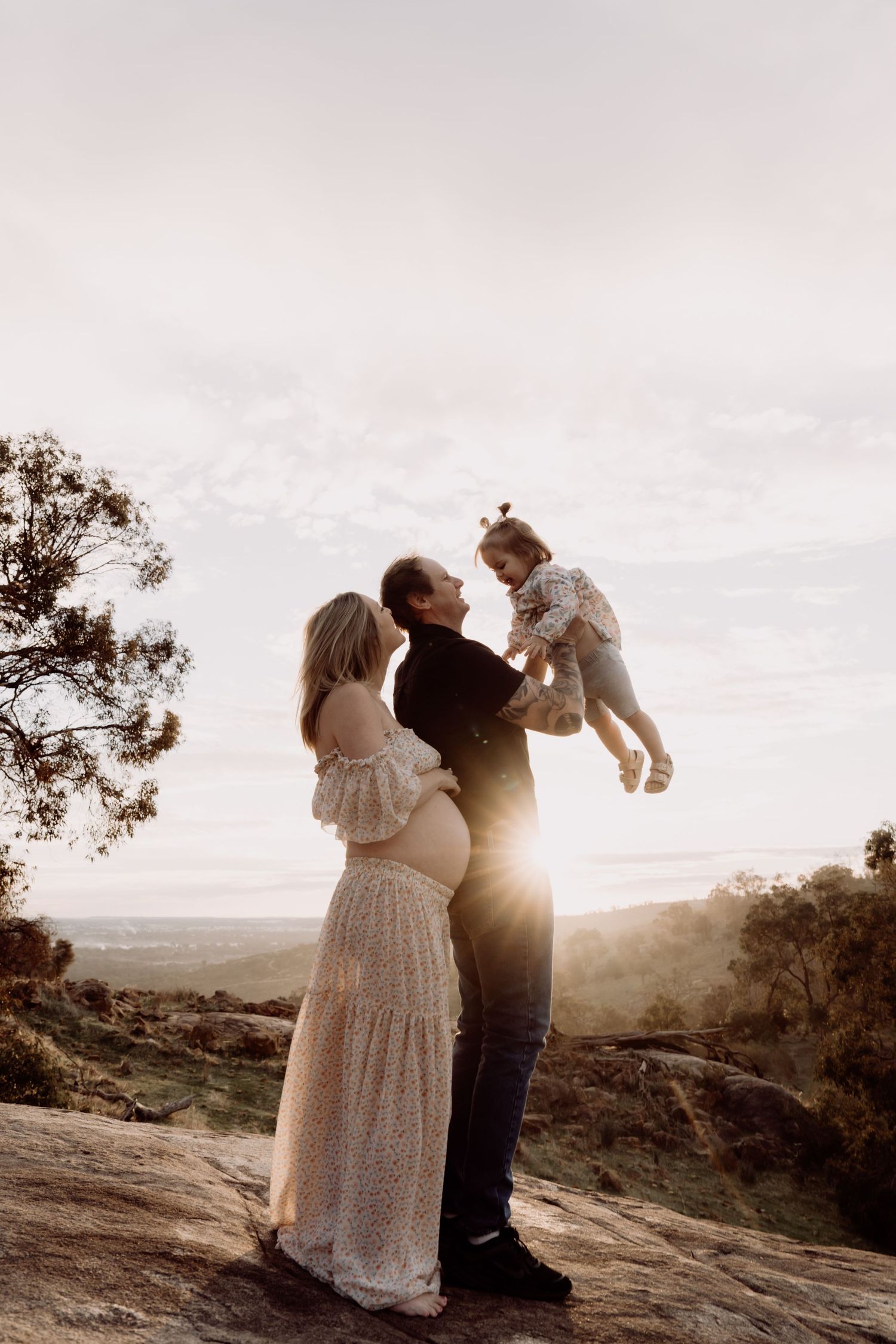 A couple stands on a rocky overlook at sunset while lifting a small child against a scenic mountain backdrop.