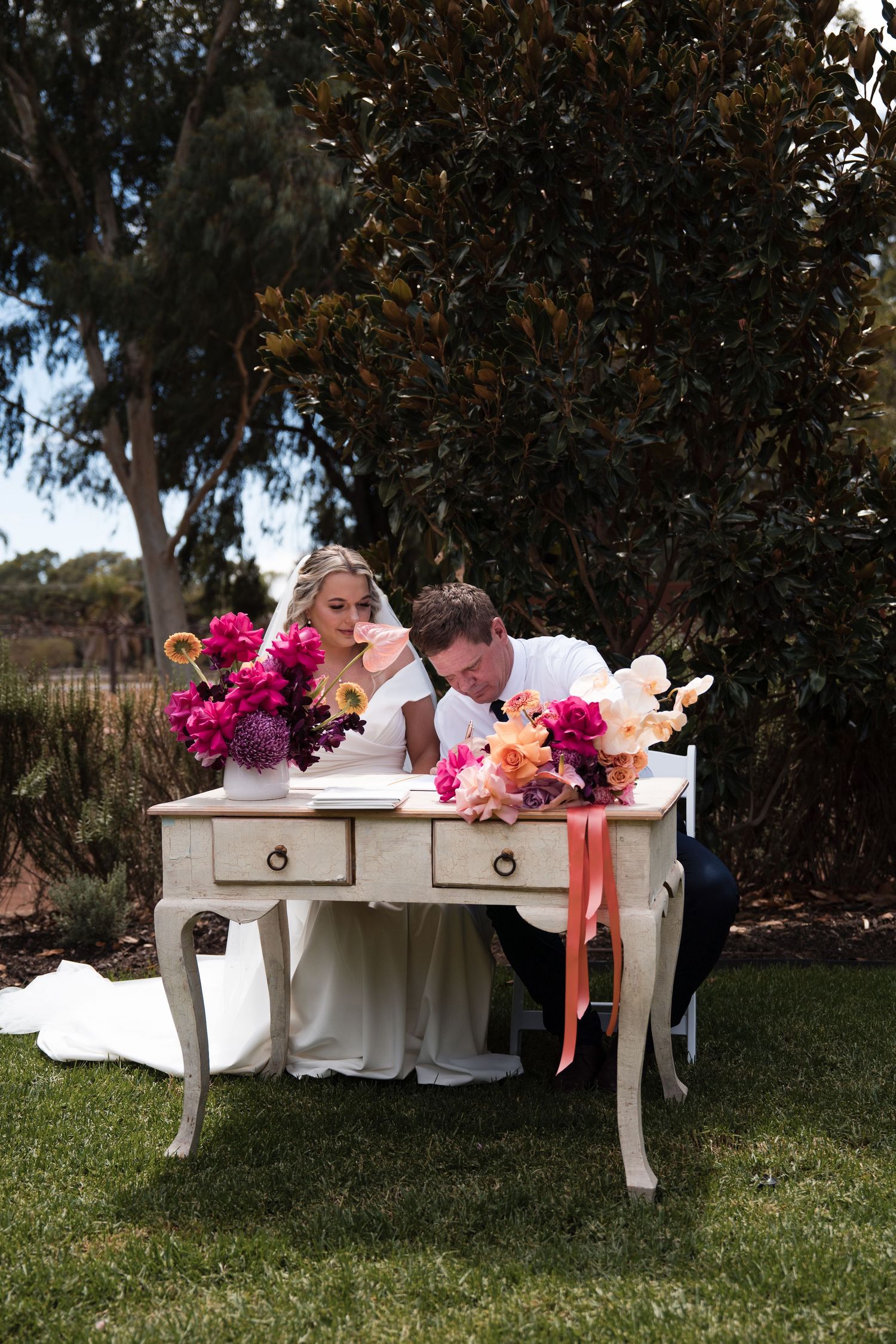 A white vintage desk adorned with vibrant pink and coral flowers sits on a grassy lawn during an outdoor event.