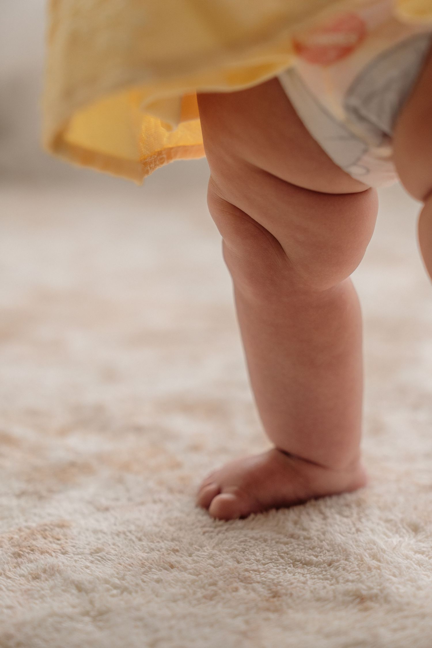Close-up view of a baby's chubby leg and foot standing on plush beige carpet with a yellow garment visible above.