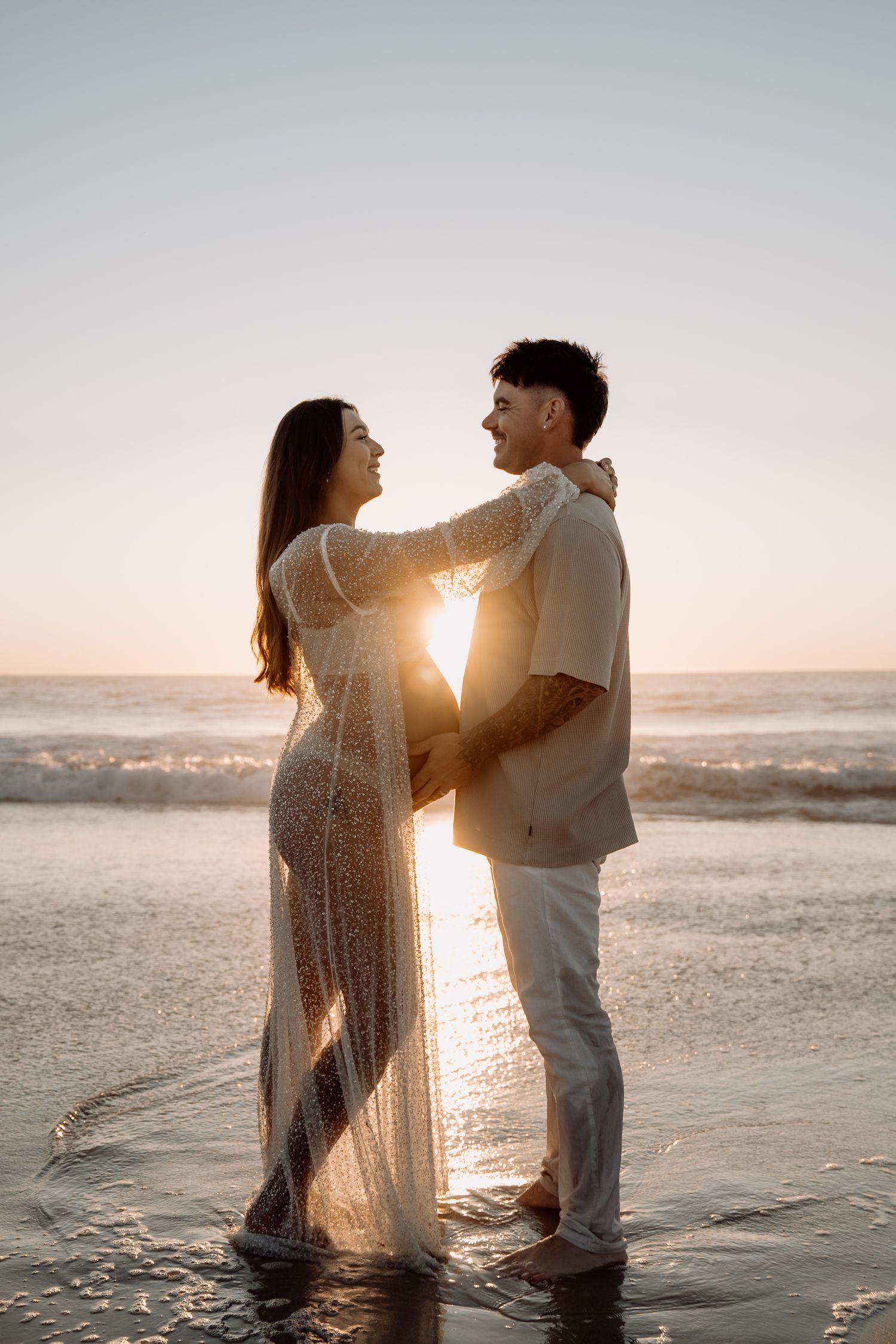 A couple stands in shallow ocean water at sunset, sharing intimate moments as golden light streams through their silhouettes.