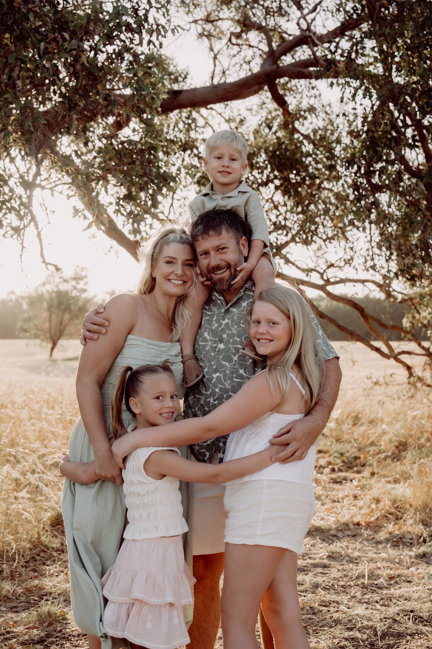 A family wearing casual summer outfits poses together in a sun-drenched outdoor setting with trees in the background.