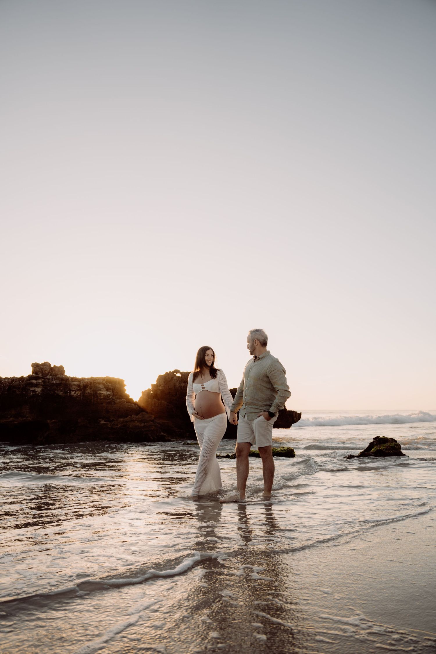 Pregnant couple standing in ocean waves at sunset on a rocky beach, holding hands in neutral tones.