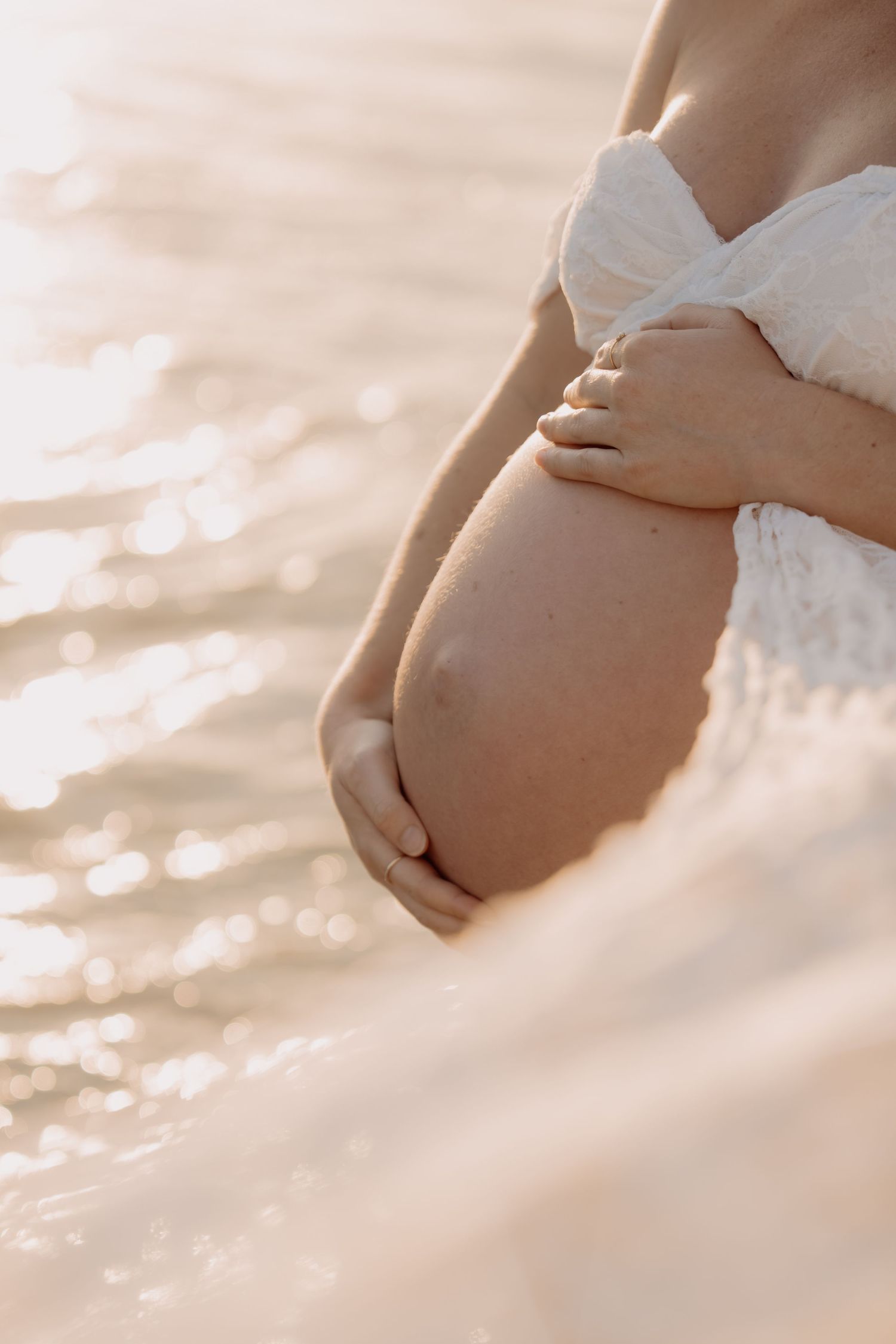 Pregnant woman in white top cradling baby bump near shimmering golden water at sunset.