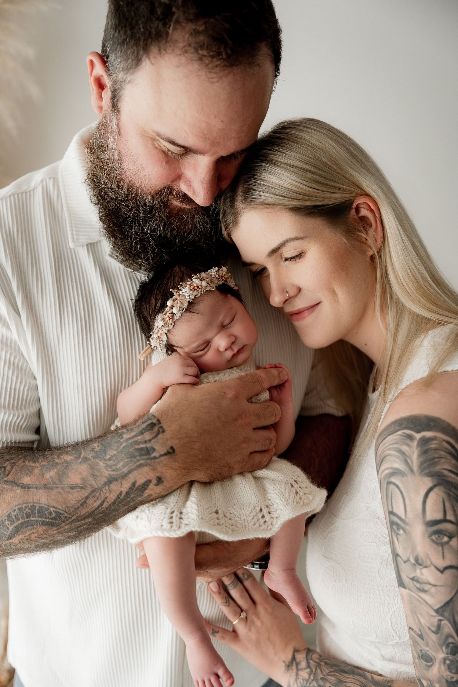 Tattooed parents tenderly holding their sleeping newborn baby girl wearing a floral headband in white outfits.
