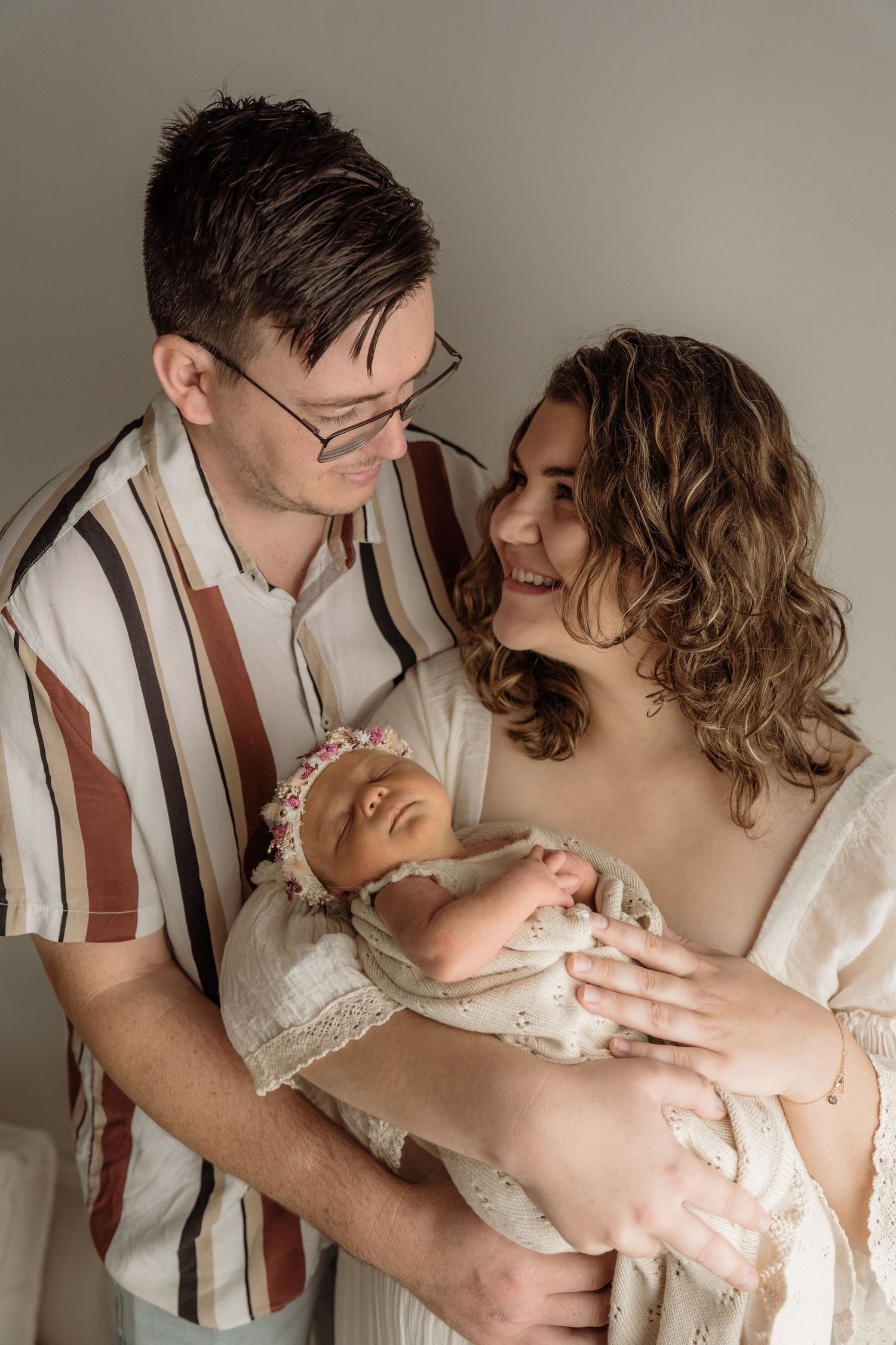 A loving couple holds their newborn baby during an intimate indoor photo session with soft neutral colors.