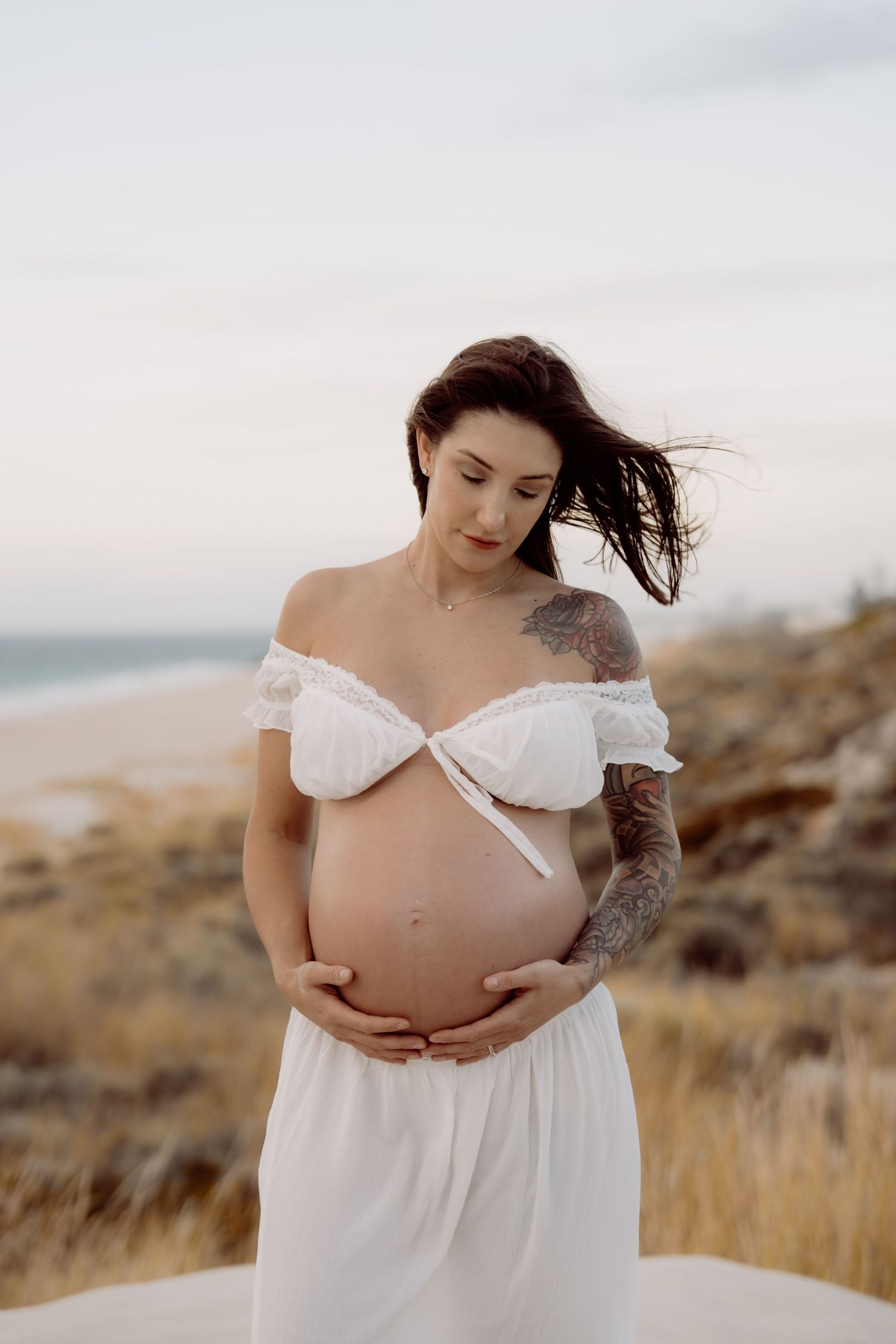 A maternity photoshoot shows a person in a flowing white dress cradling their pregnant belly on a grassy hillside.