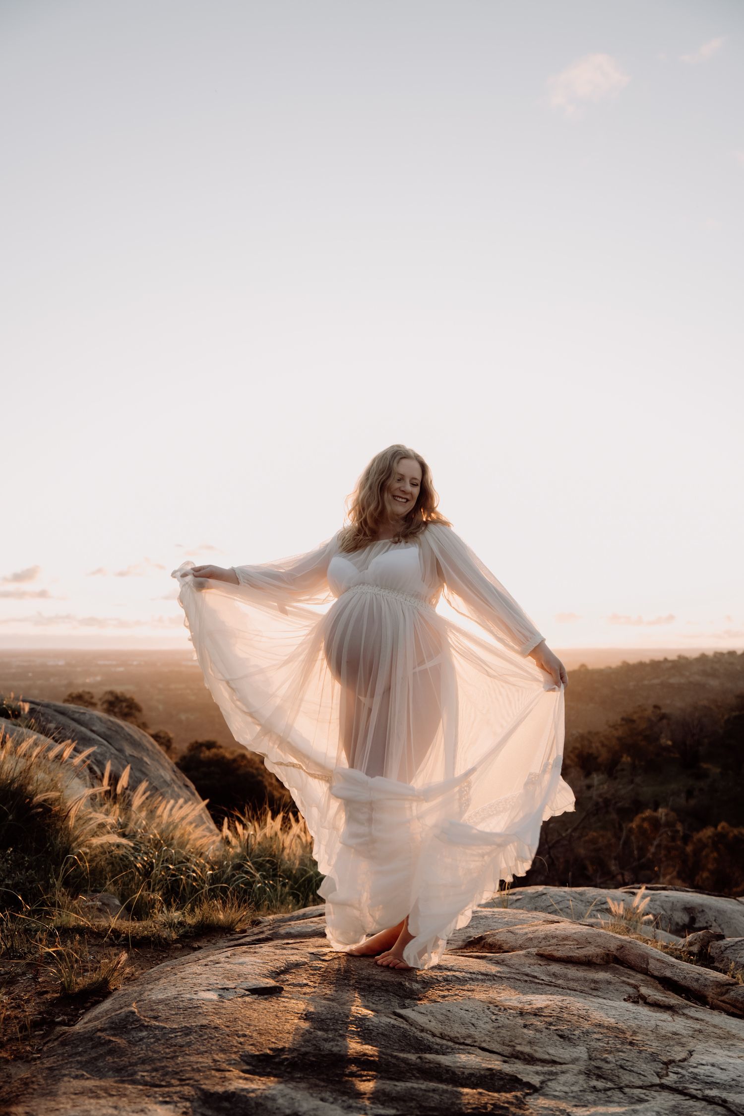 A flowing white dress moves gracefully in the wind as someone stands on rocky terrain during a warm sunset.