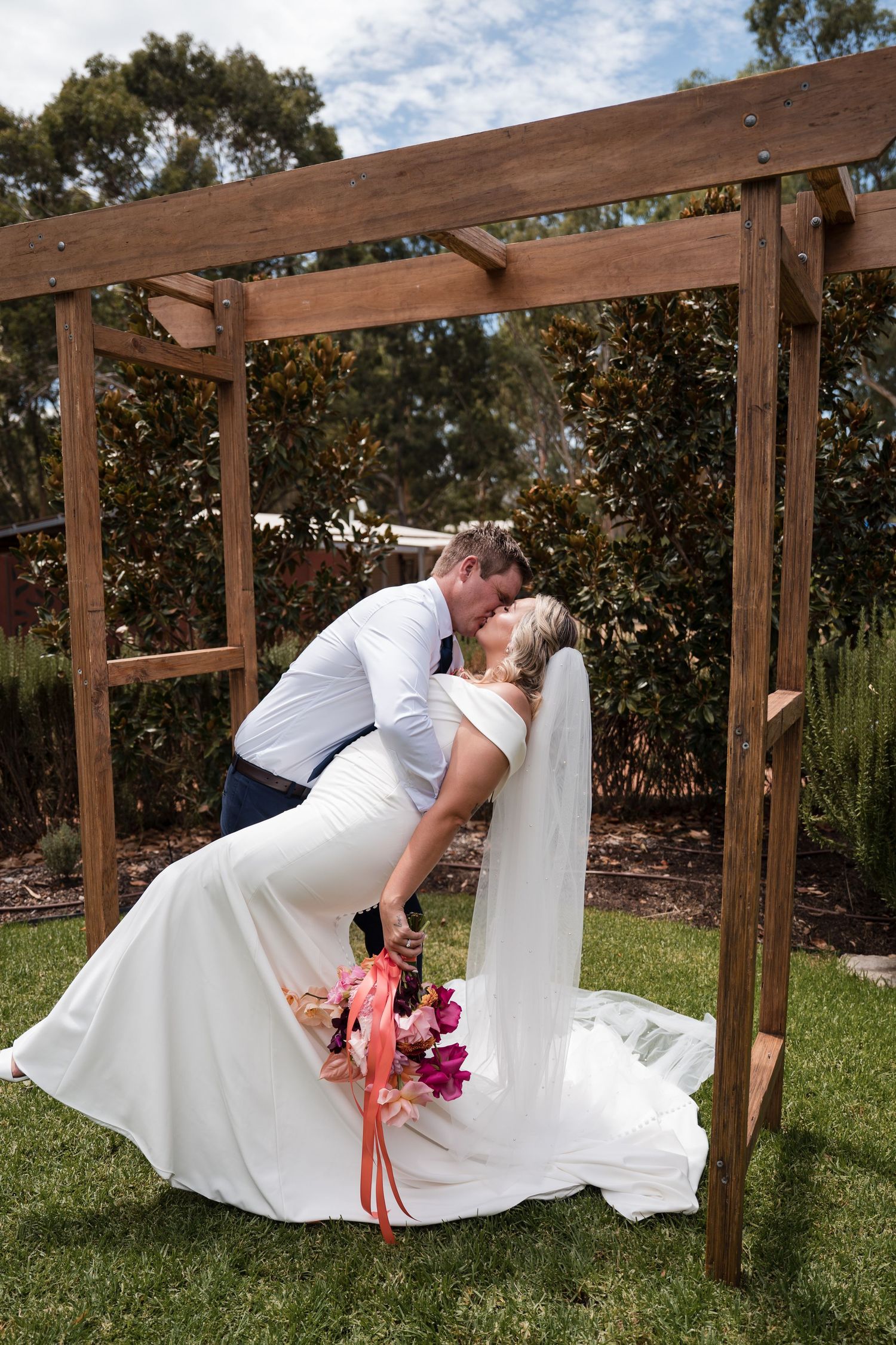 A couple shares a romantic kiss under a wooden pergola in a garden setting during their wedding celebration.