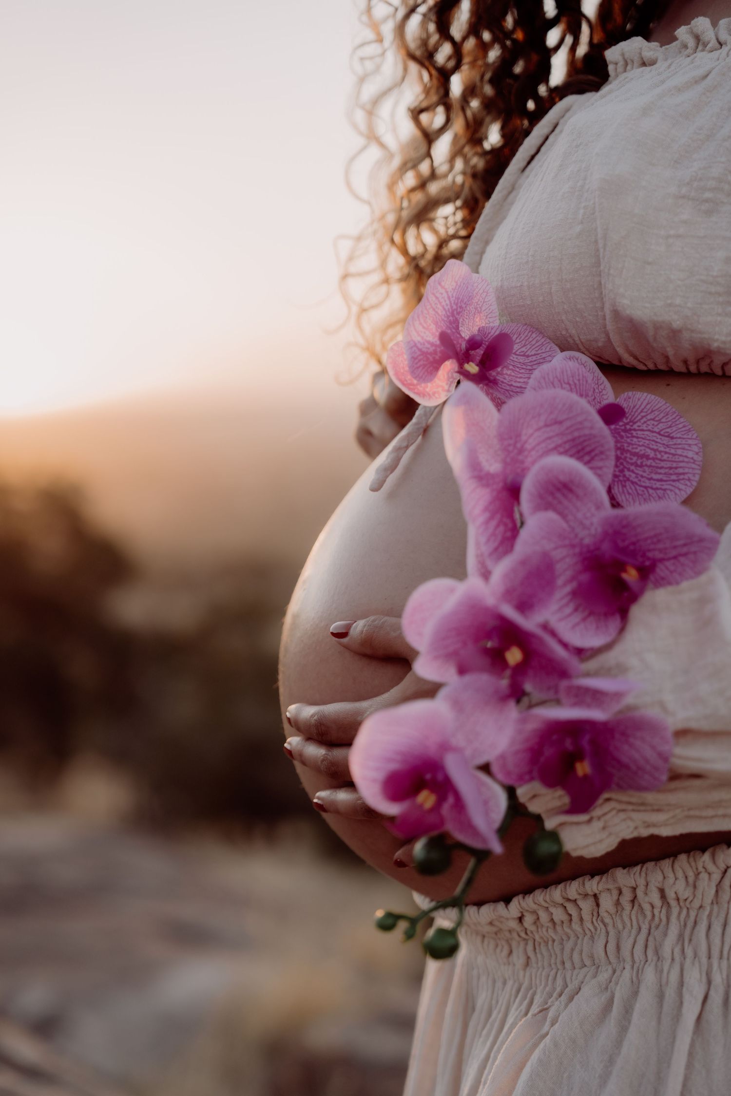 A person in a white dress holds delicate pink orchids against a sunset backdrop.