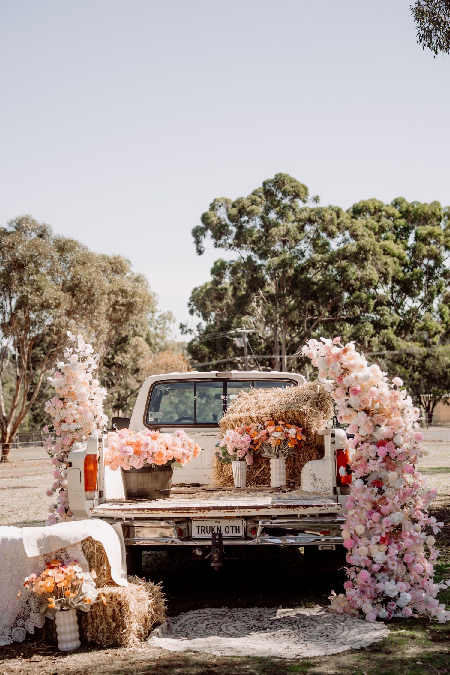 White pickup truck decorated with pink floral arches and hay bales at an outdoor rustic wedding celebration.