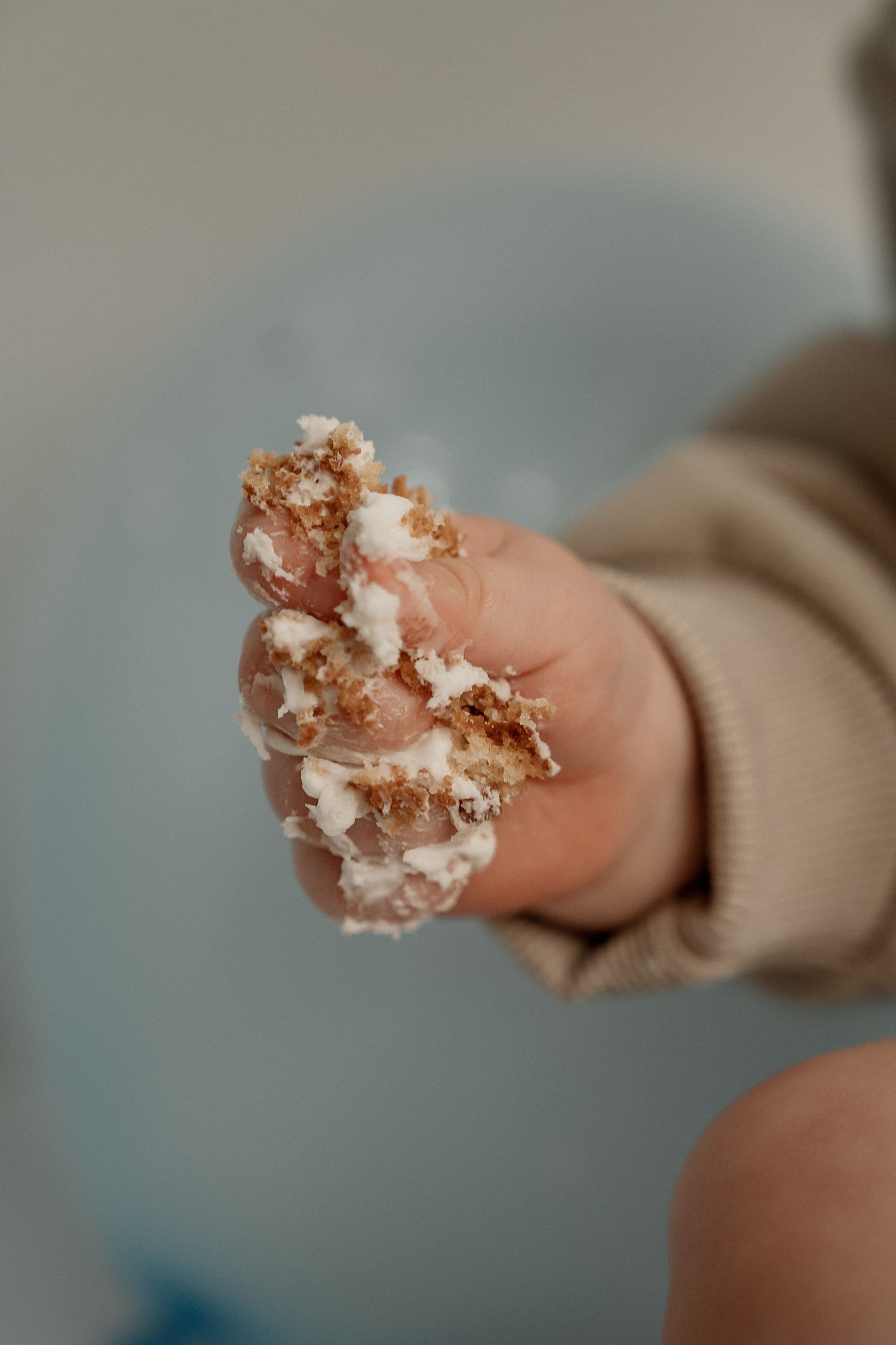 A small hand holds a crumbled piece of cake or cookie with white frosting against a blurred blue background.
